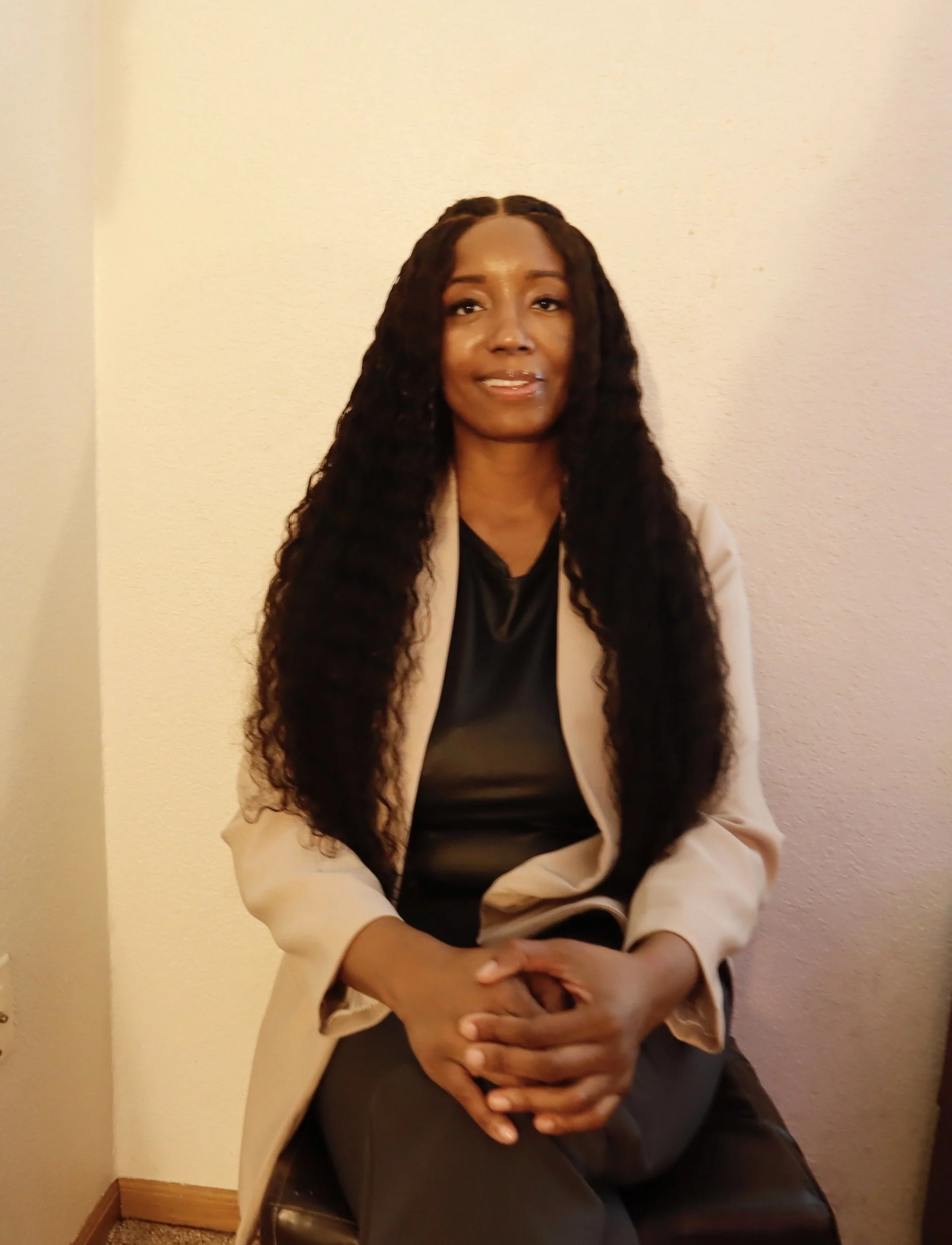 A woman sitting on a chair with her hands clasped on her lap, wearing a black top and a beige blazer, long curly dark hair, and posing against a plain light-colored wall.