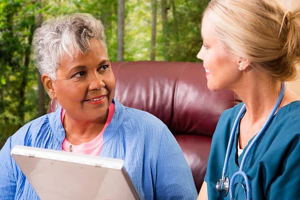 An elderly woman and a nurse are sitting and talking indoors, with a blurred green outdoor background visible through a window.