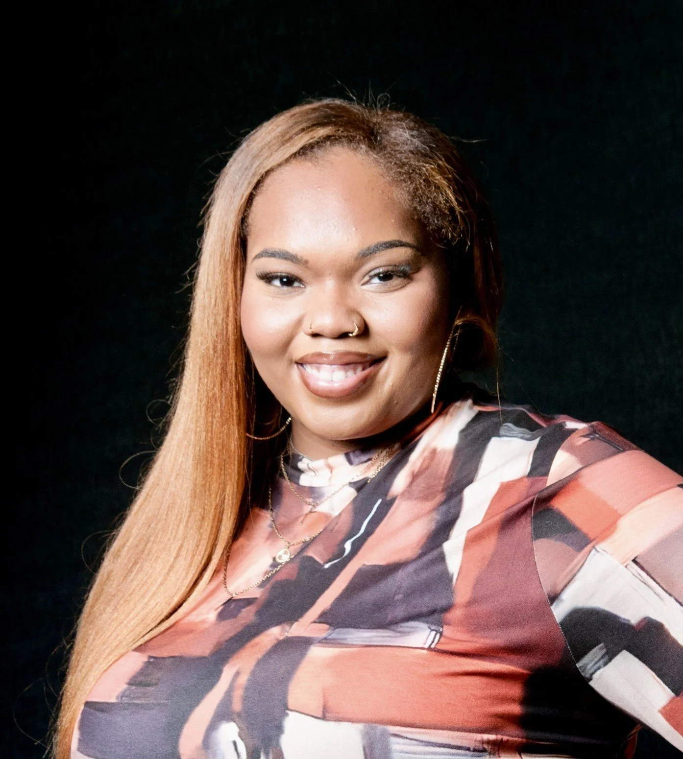 Portrait of a smiling woman with long blonde hair, gold hoop earrings, and layered necklaces, wearing a colorful abstract patterned top, against a black background.