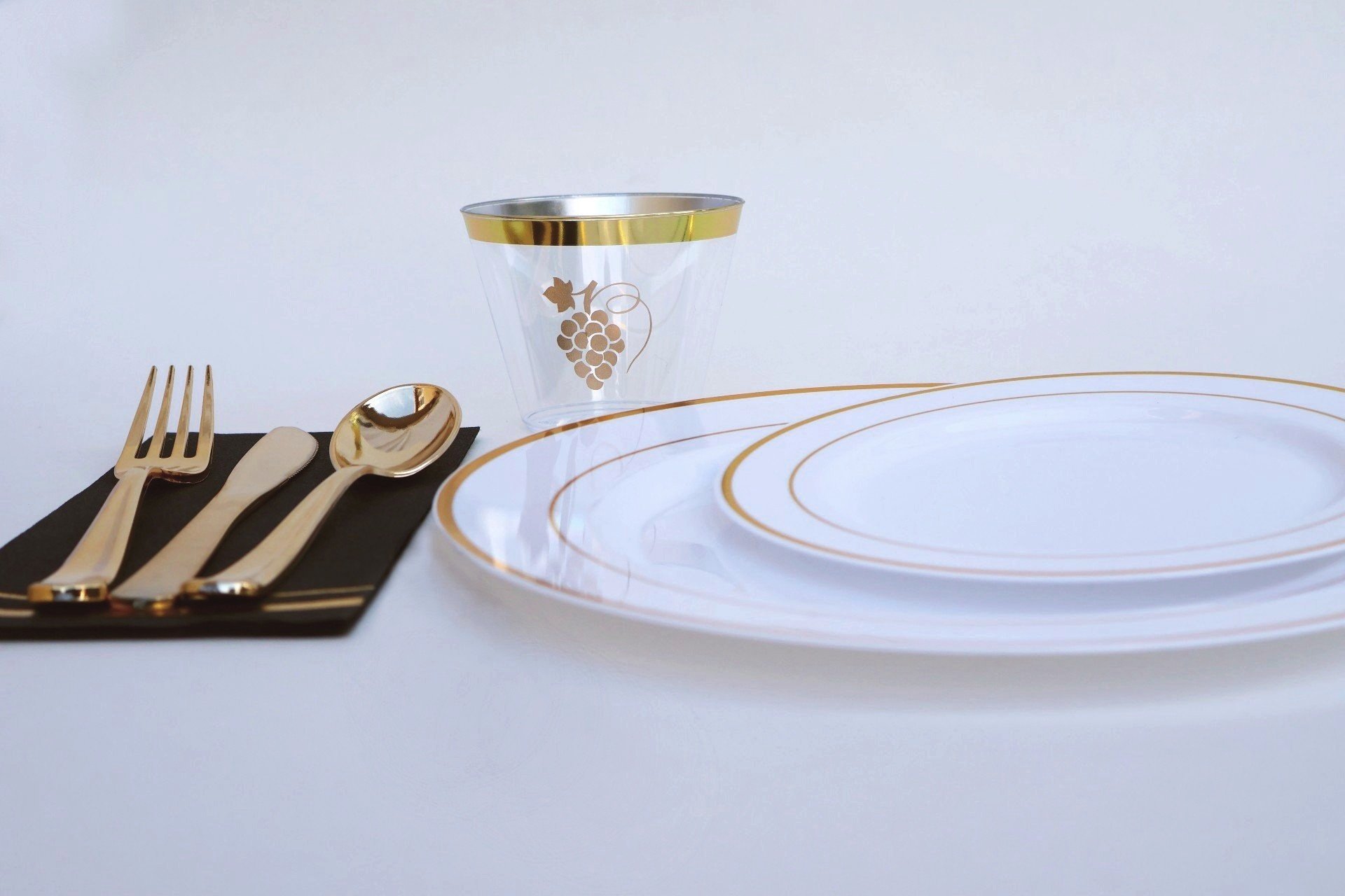 table setting with two gold and white plates, clear glass with gold trim and gold grape bunch logo, black napkin with gold silverware on top