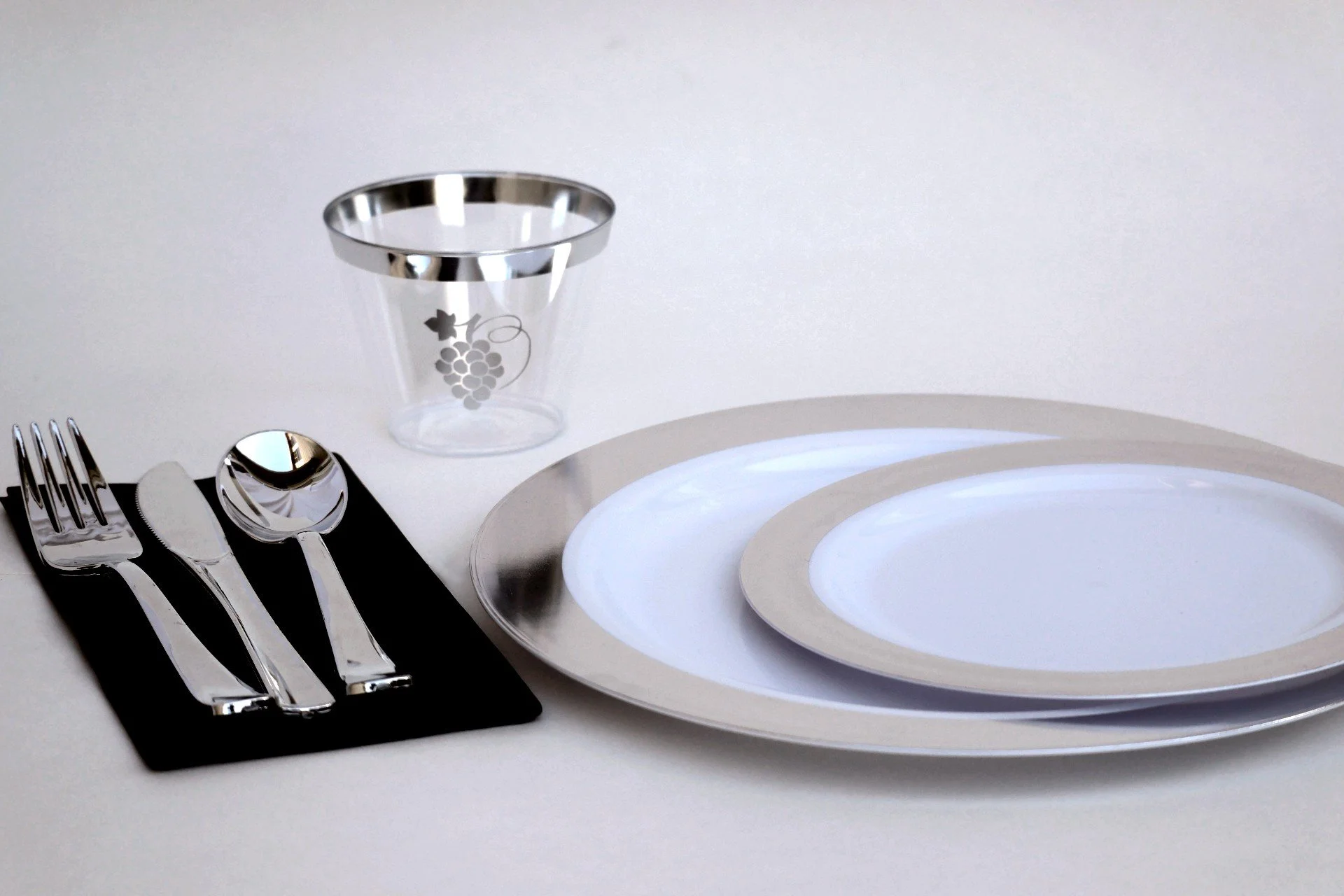 table setting with two white and silver plates, clear class with silver trim and silver grape bunch logo, black napkin and silver silverware on top