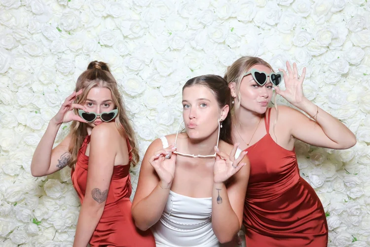 Bride and bridesmaids in heart sunglasses at a white rose flower wall photo booth at a Winnipeg wedding