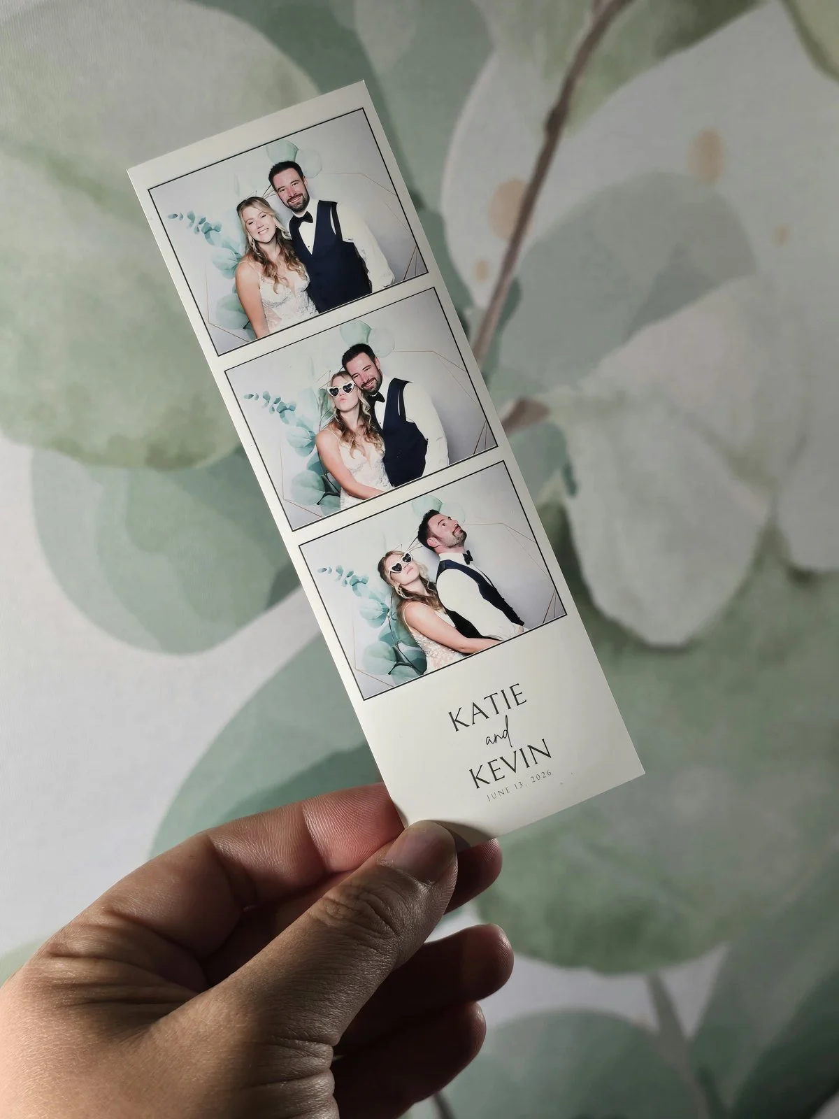 A hand holding a wedding photo strip with the couple's names and date printed at the bottom