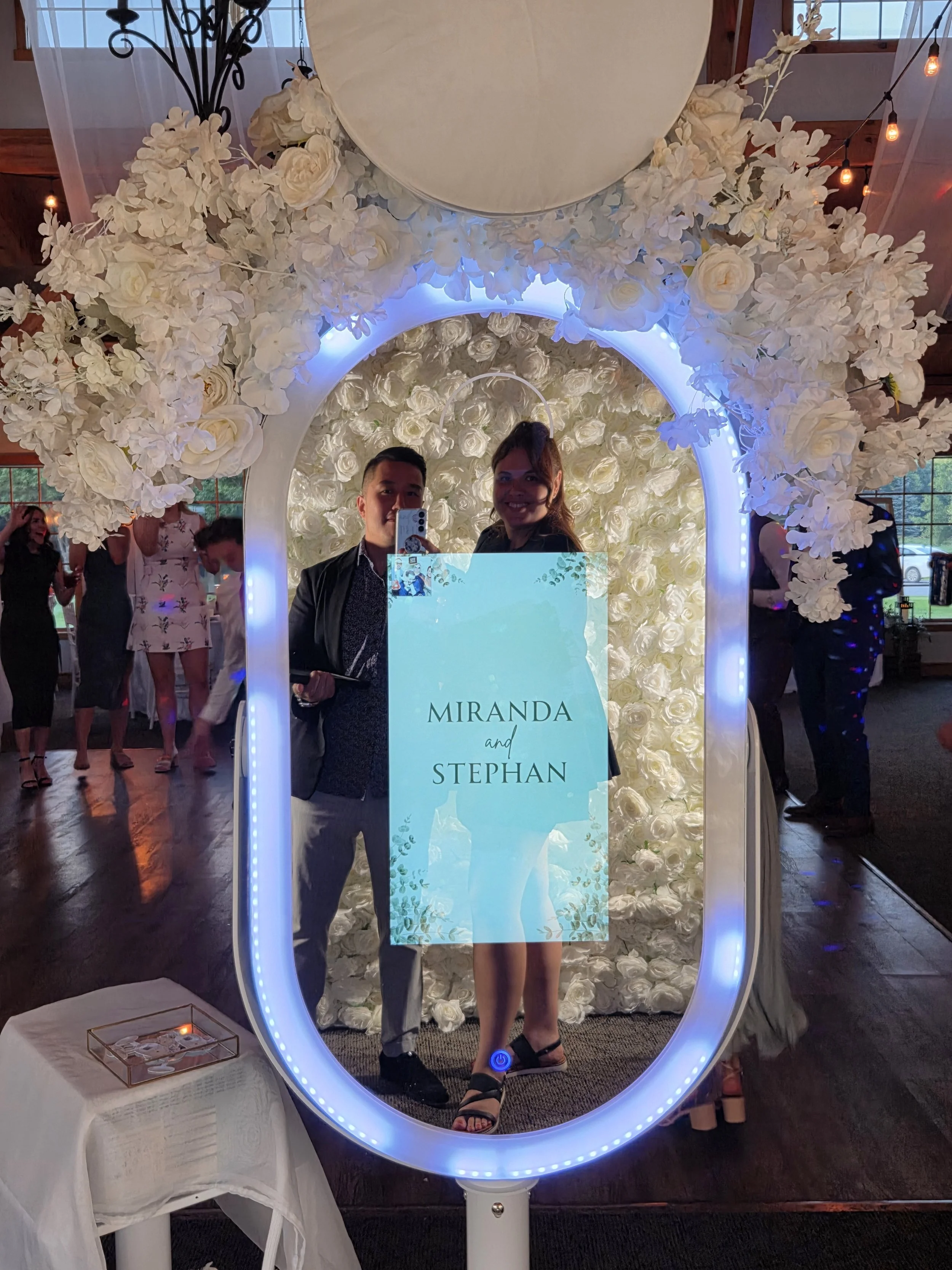 Large oval mirror photo booth with led lights and white flowers on top. A couple is posing in the mirror.