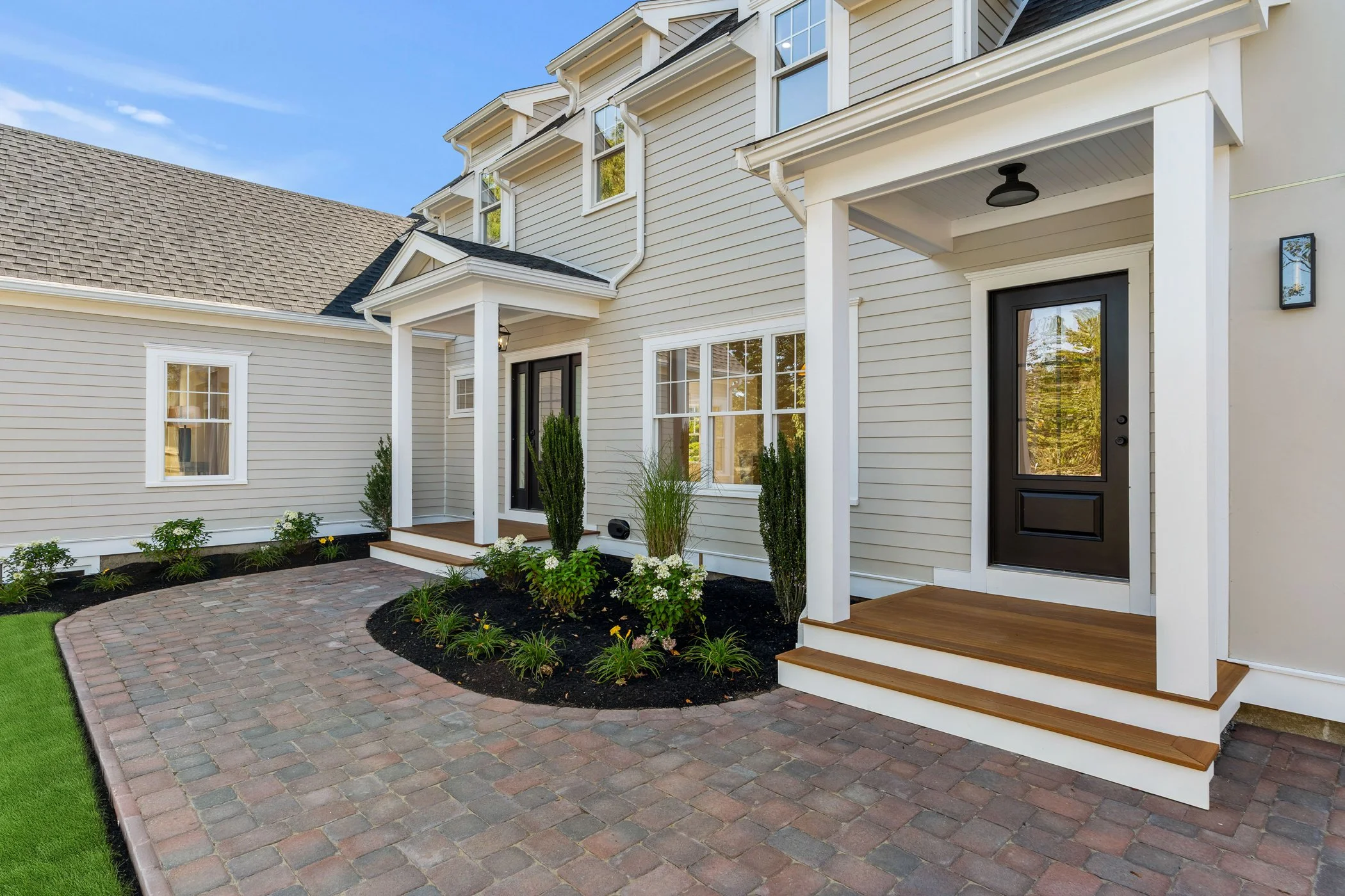 Front view of a modern house with beige siding, black front door, porch steps, garden with plants, and brick walkway under a blue sky.