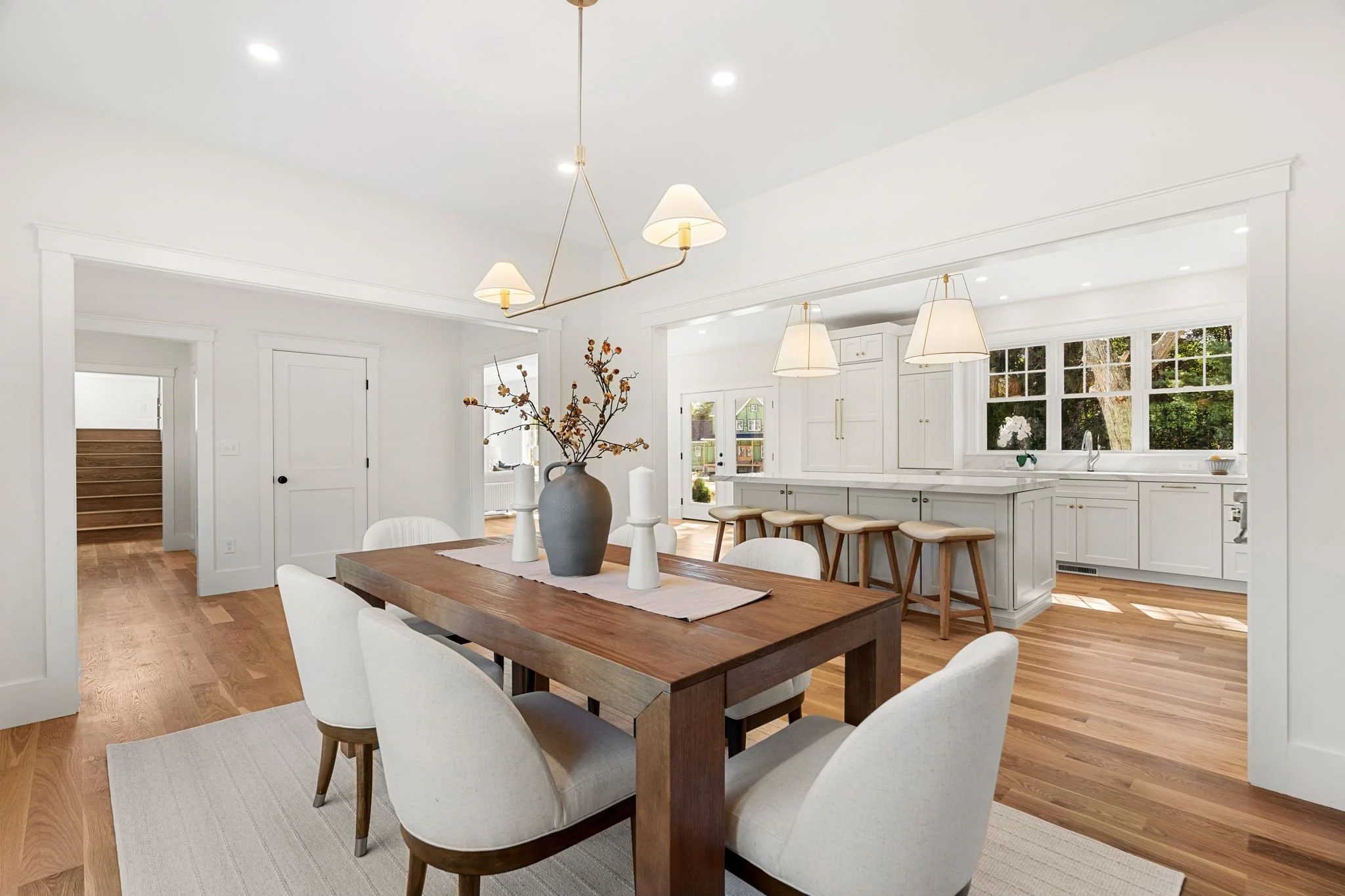 Bright, open-concept kitchen and dining area with white walls, large windows, and hardwood floors. Centered is a wooden dining table with white upholstered chairs and a large vase with branches as a centerpiece. Overhead lighting includes a modern chandelier and pendant lamps.