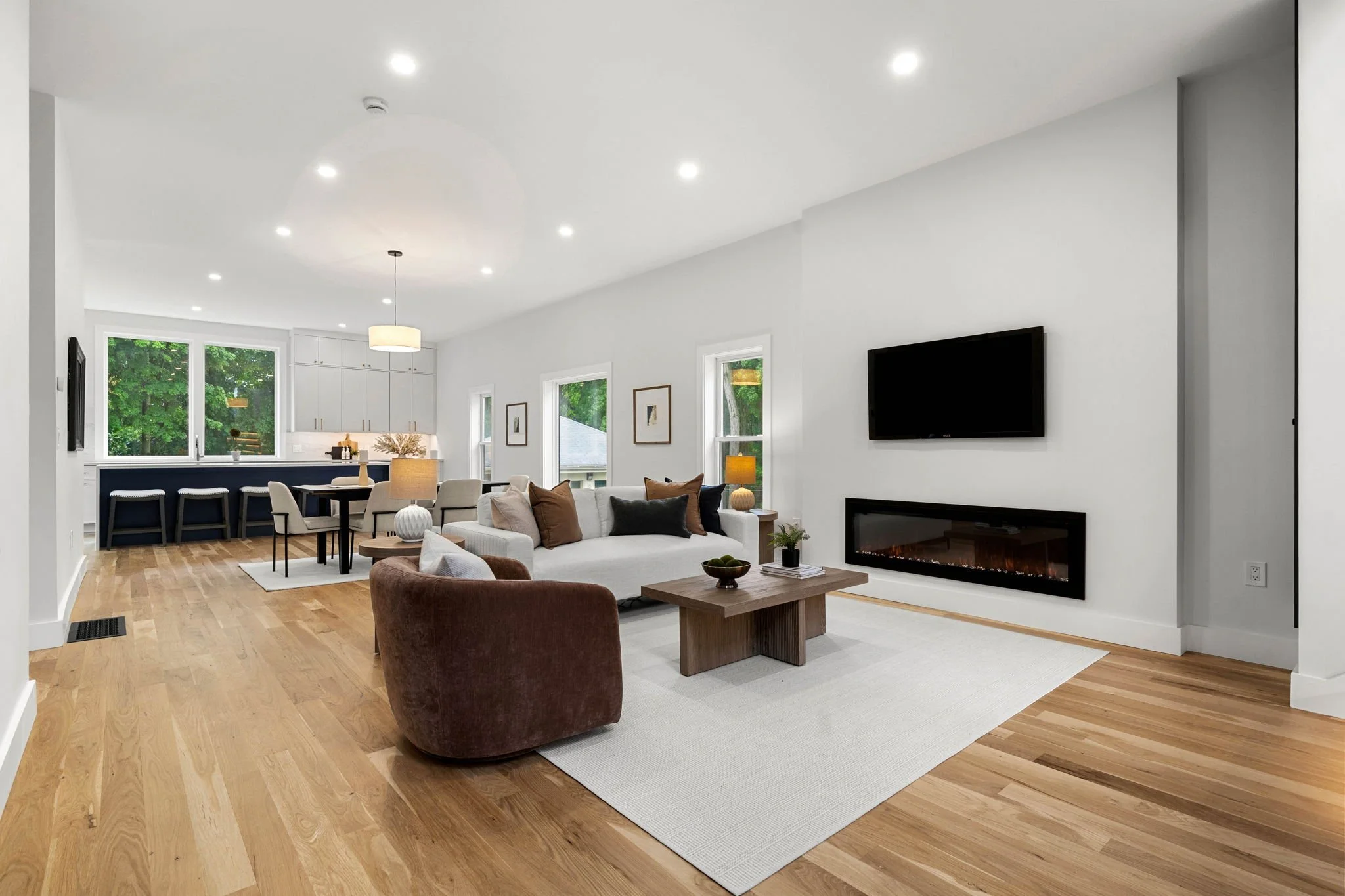 A modern, open-concept living space with white walls, hardwood floors, a mounted flat-screen TV, a fireplace, a white sofa with brown cushions, a coffee table with books and plants, a brown armchair, and large windows showing greenery outside.