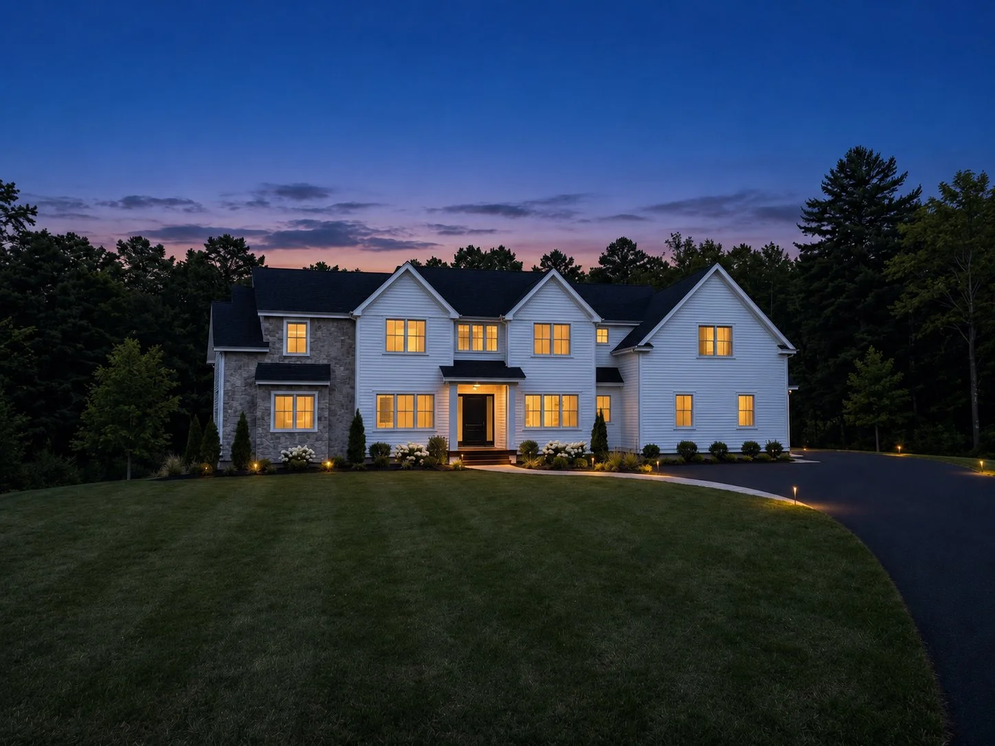 Large modern house with white siding and stone accents, illuminated windows, surrounded by landscaped yard and trees at dusk.