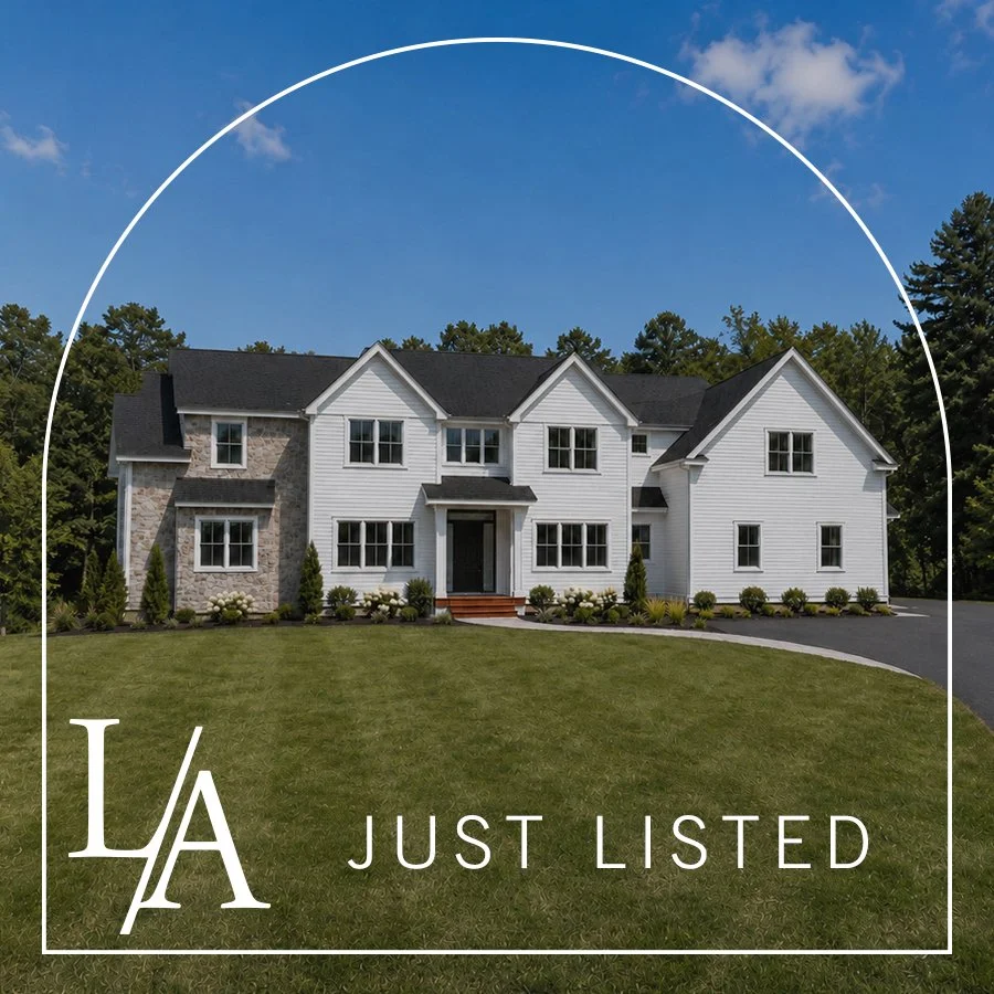 A large two-story house with white siding, gray stone accents, and a black roof. The house has multiple windows, a small front porch with stairs, and is surrounded by a well-maintained lawn and landscaped bushes. The background shows a blue sky with some clouds and trees. The overlay text says "LA JUST LISTED."