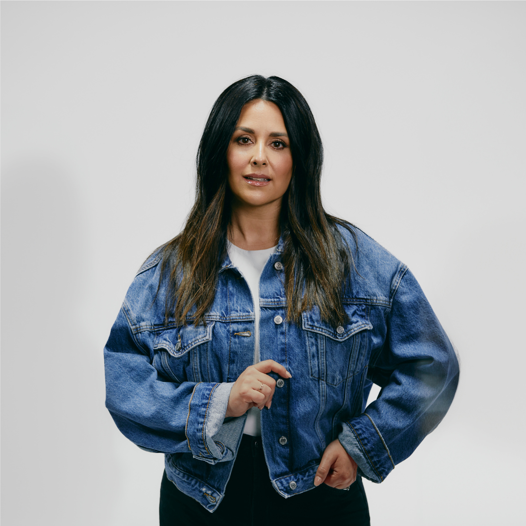 A woman with long dark hair wearing a denim jacket, white shirt, and black pants standing against a plain background.