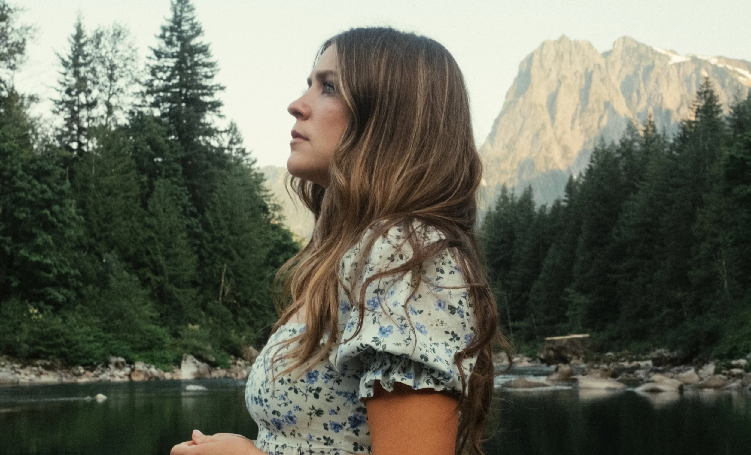 A woman standing by a river with forest and mountain in the background