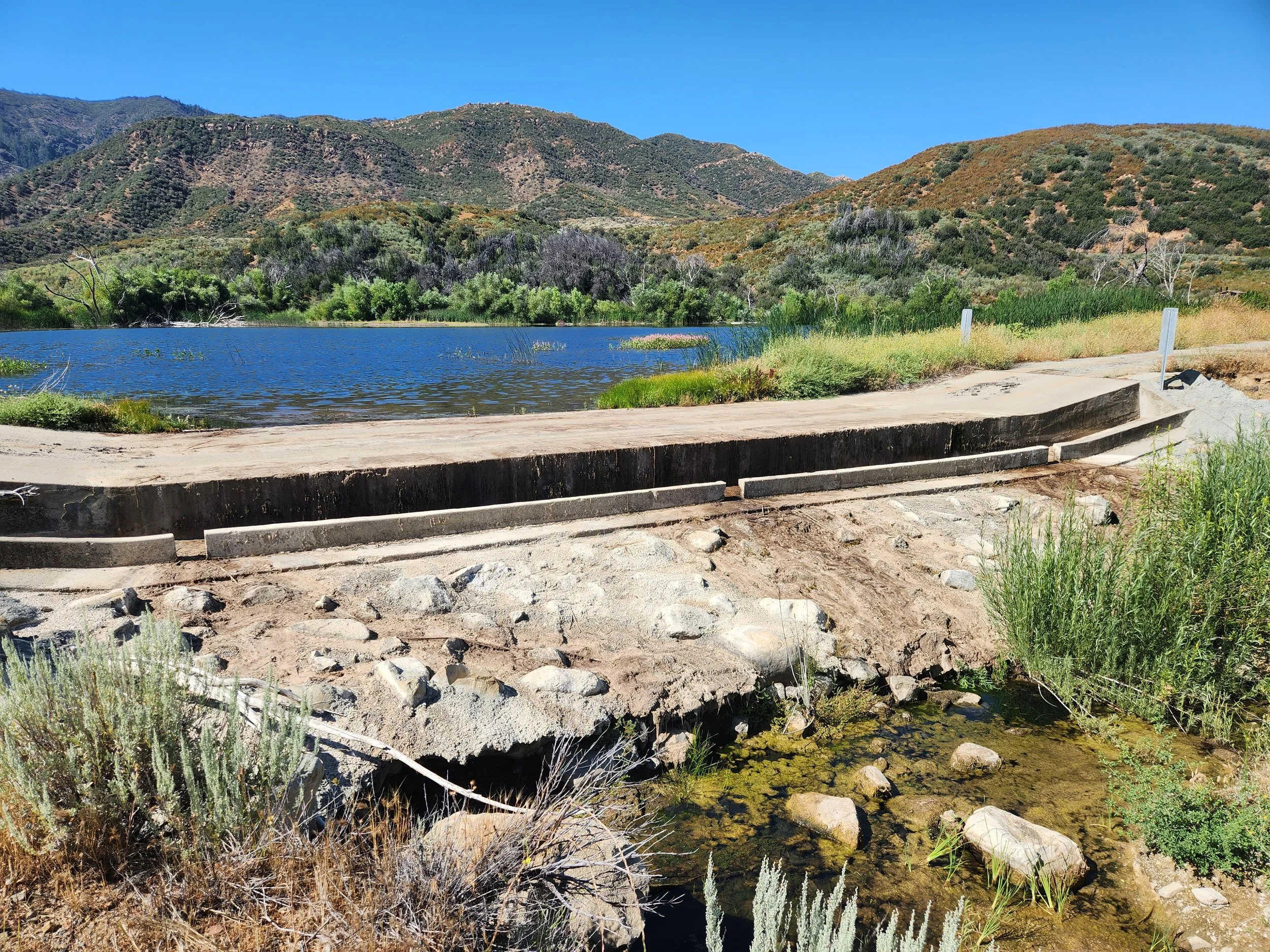 Concrete structure by a small body of water with mountains and vegetation in the background.