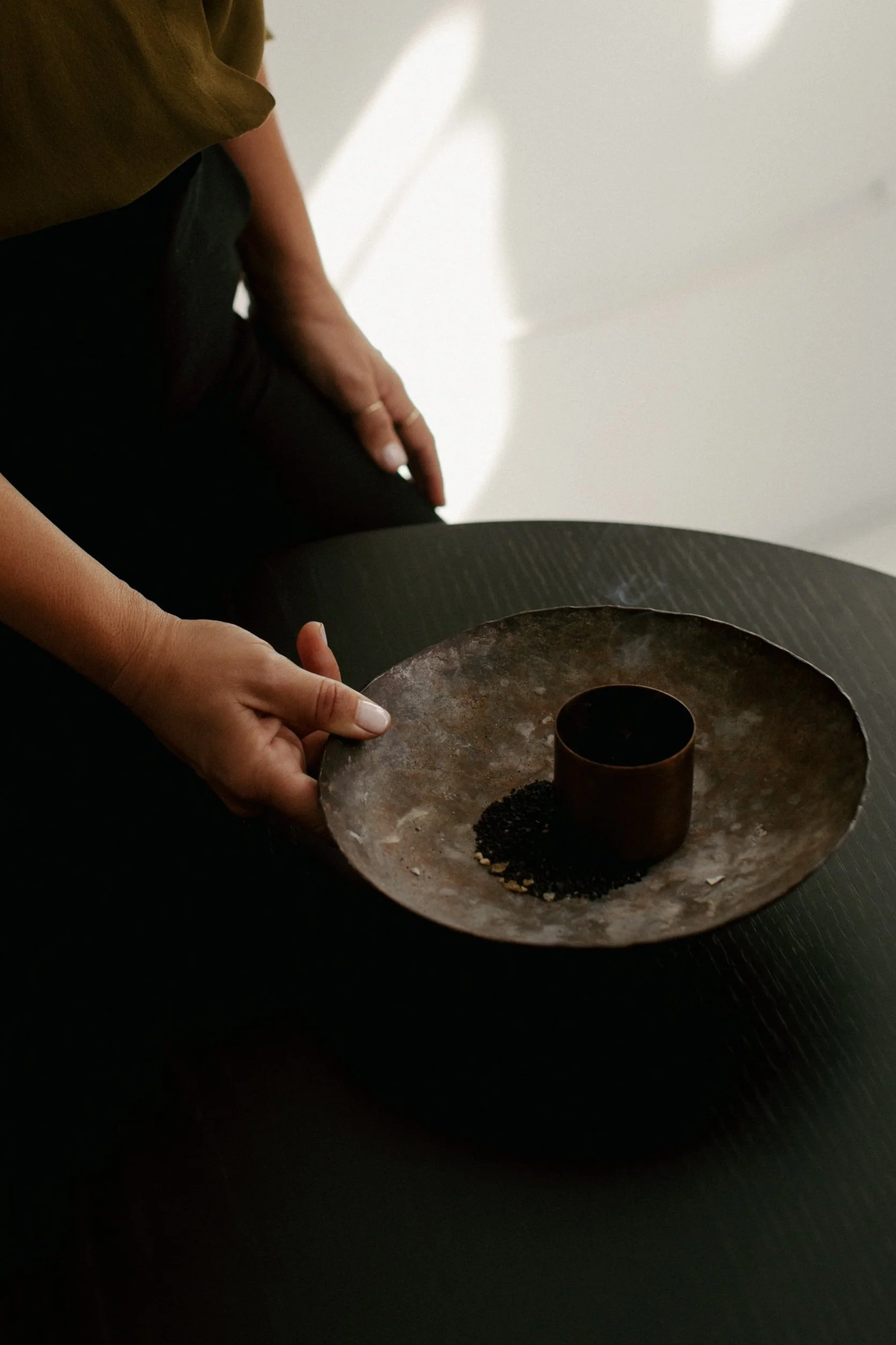 Person holding a round, metallic tray with a cylindrical cup and black granules or seeds on it, sitting at a dark table.