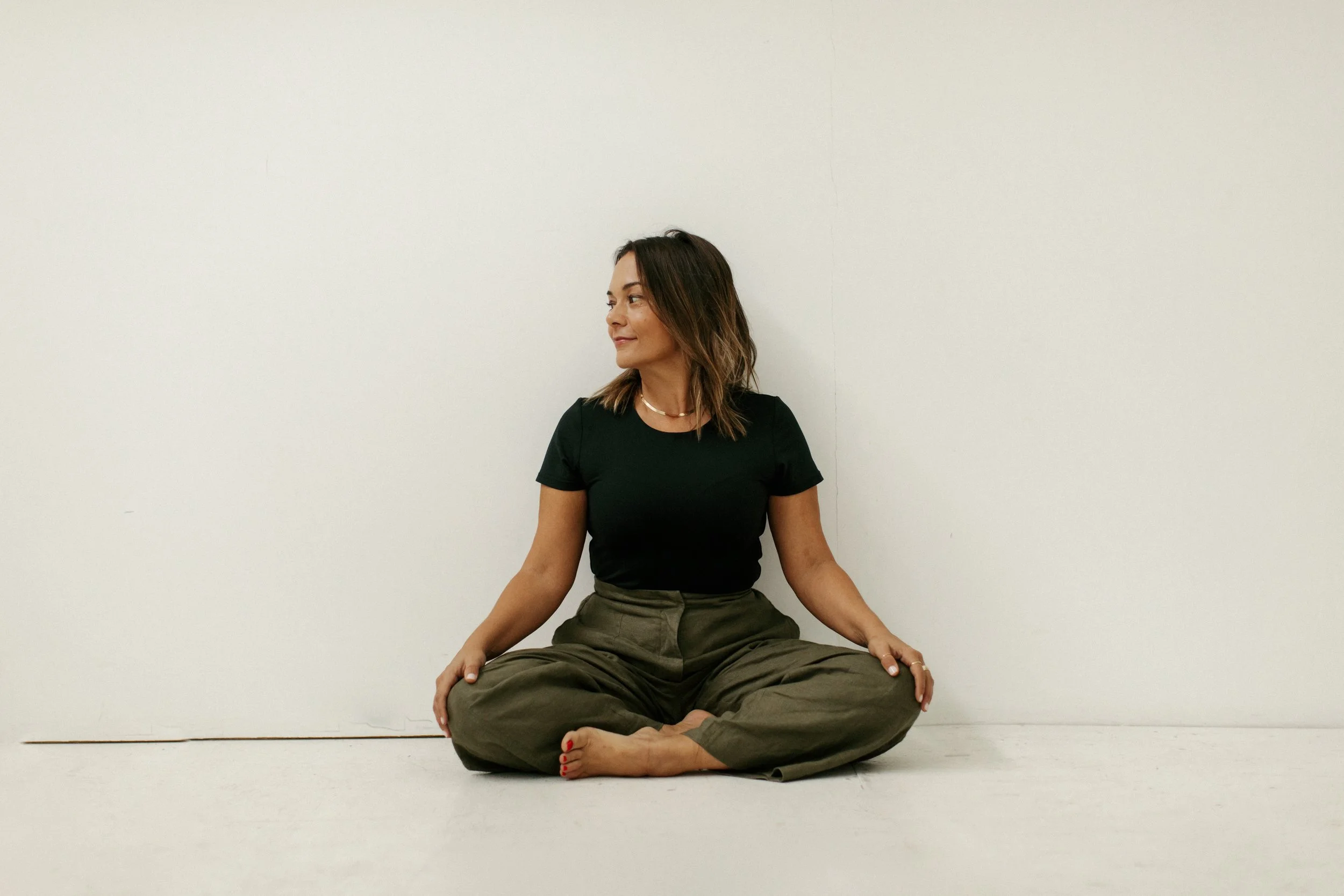 Woman sitting cross-legged on the floor against a plain white wall, wearing a black t-shirt and khaki pants.