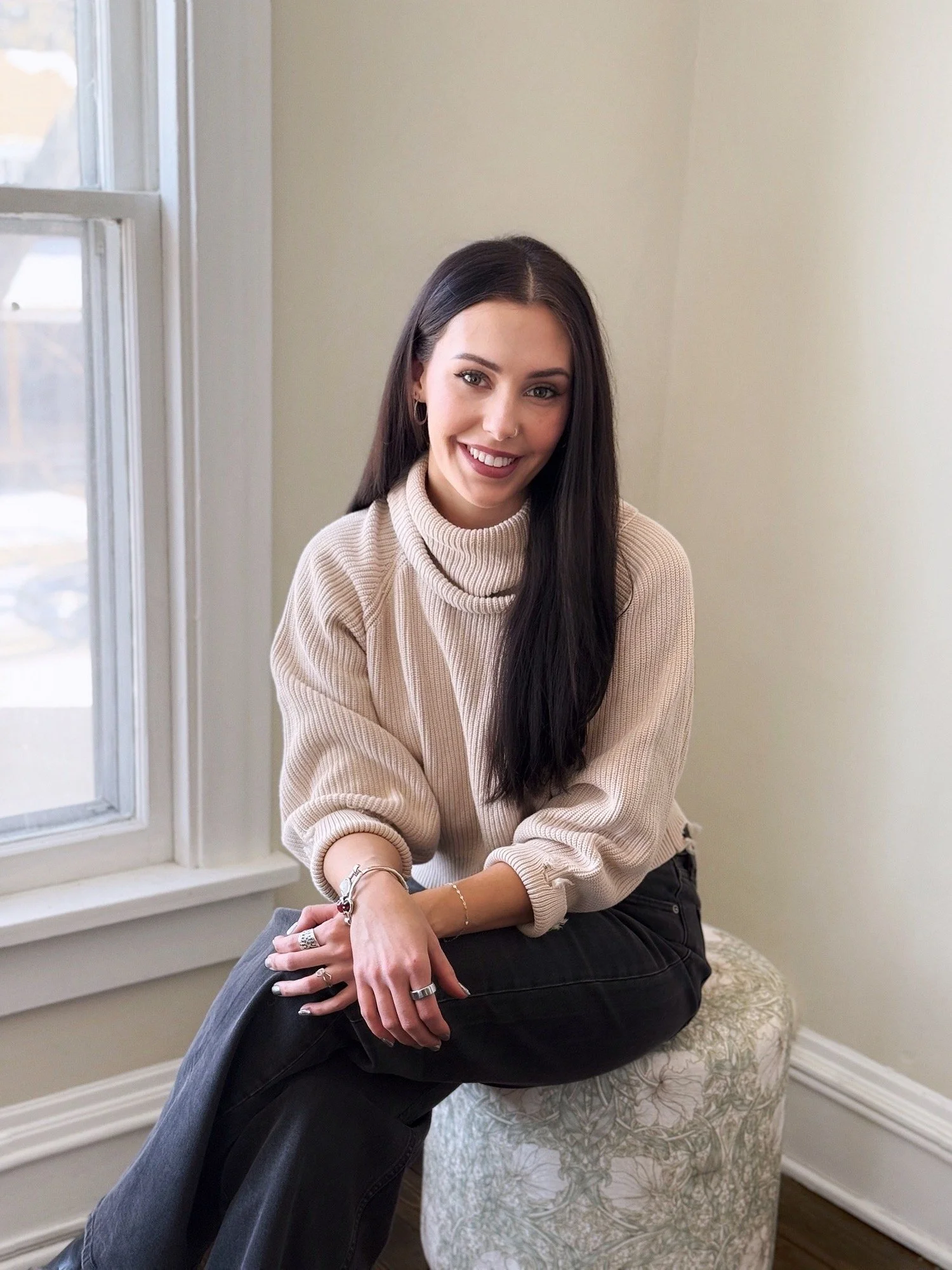 Jasmyn Stengel, LMFT-IT, sits near a window with her legs and hands crossed. She wears a beige turtle neck, and black pants. Long brunette hair drapes over her shoulder.