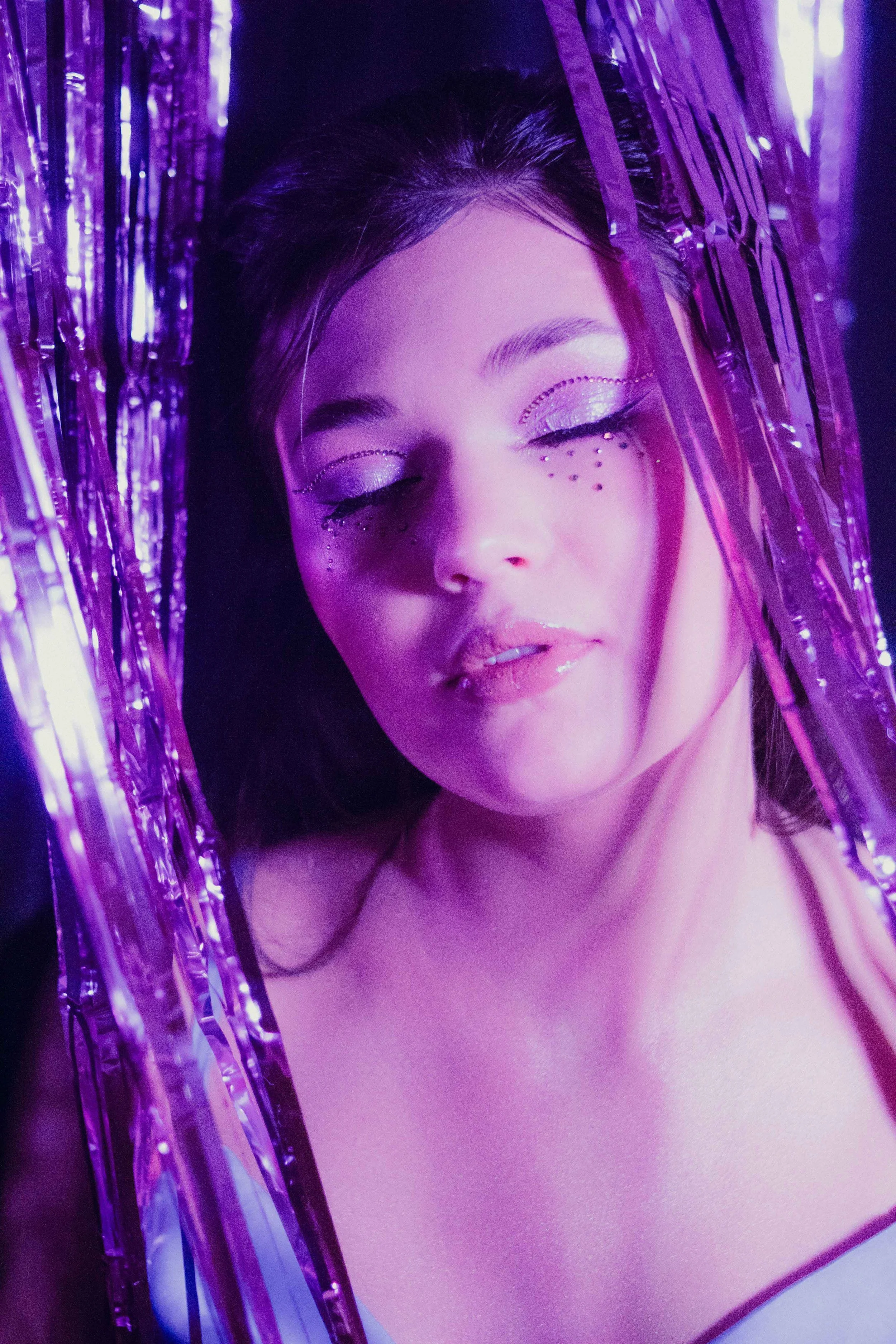 Editorial beauty photo of a woman framed by purple metallic fringe with eyes closed, featuring sparkling eyeshadow and face crystals under purple lighting