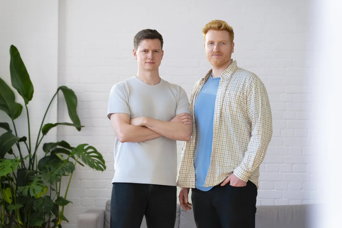 Two men standing indoors against a white brick wall with a large green plant on the left. One man has arms crossed and wears a light gray t-shirt, while the other has his hand in his pocket and wears a blue t-shirt with an open plaid shirt over it.