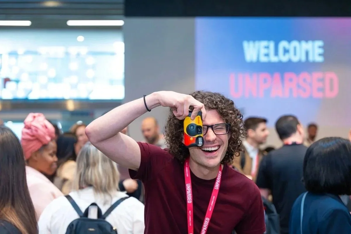 Person with curly hair and glasses laughing while taking a photo at a crowded event with a "Welcome Unparased" sign in the background.