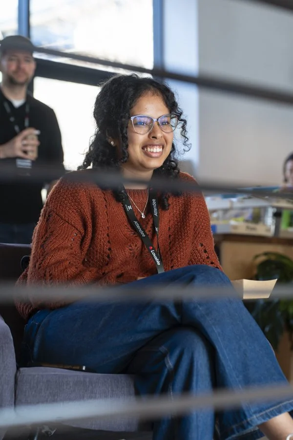 A woman with curly black hair, glasses, and a rust-colored sweater smiling while sitting on a couch in an indoor office setting, with a man in the background holding a drink.