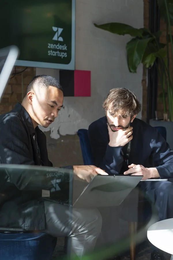 Two men discussing work at a table with a laptop in a modern office with Zendesks Startups branding on a screen in the background.