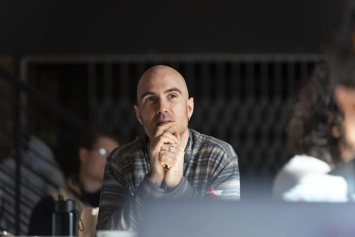 A man with a shaved head and earrings, sitting at a table in a casual setting, appears to be deep in thought with hands clasped near his mouth, talking with other people.