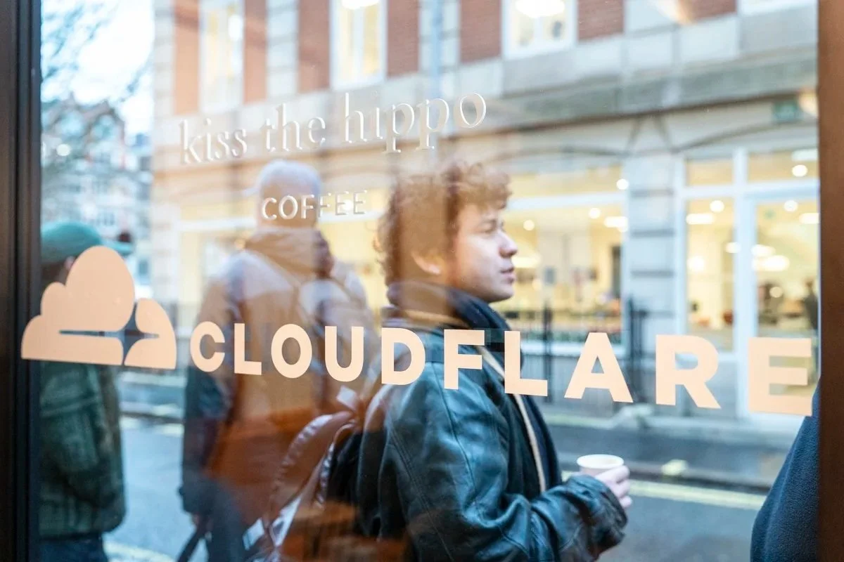 People standing outside a coffee shop with a glass window displaying the Cloudflare logo and the text 'kiss the hippo' and 'COFFEE'.