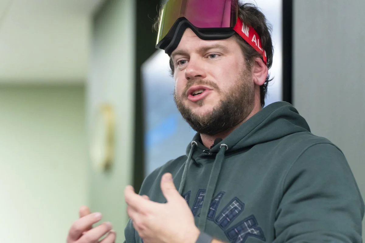 A man with a beard wearing ski goggles and a hoodie, speaking and gesturing with his hand.