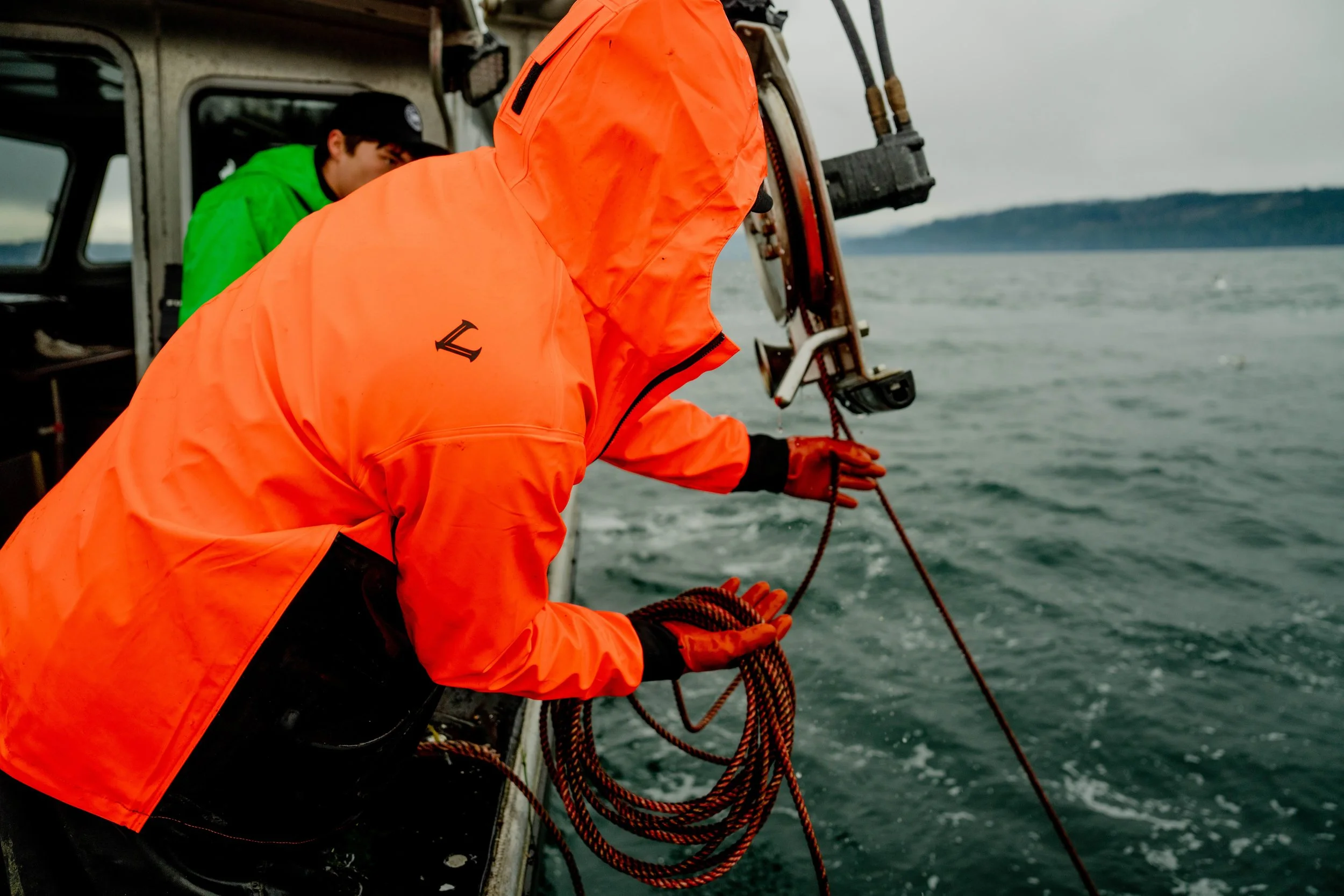 Two people on a boat, one in an orange waterproof jacket handling a rope, the other in a green jacket observing, with water and distant shoreline in the background.