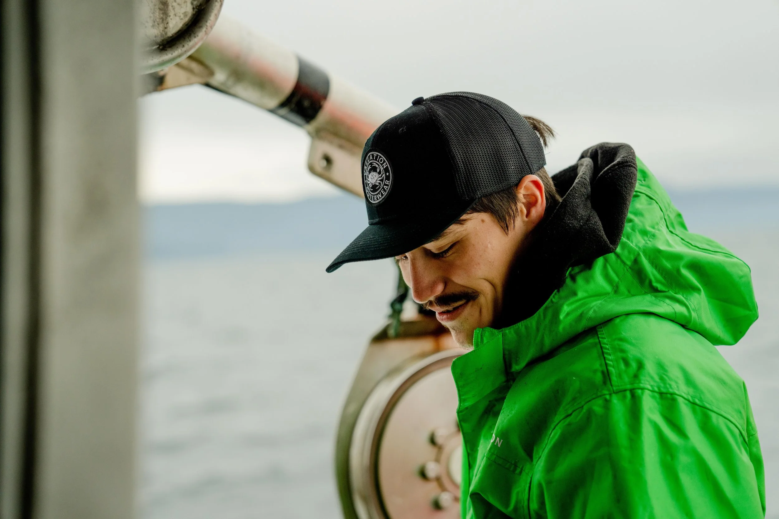 A man in a green jacket and a black hat is smiling and looking down, possibly on a boat or near water.
