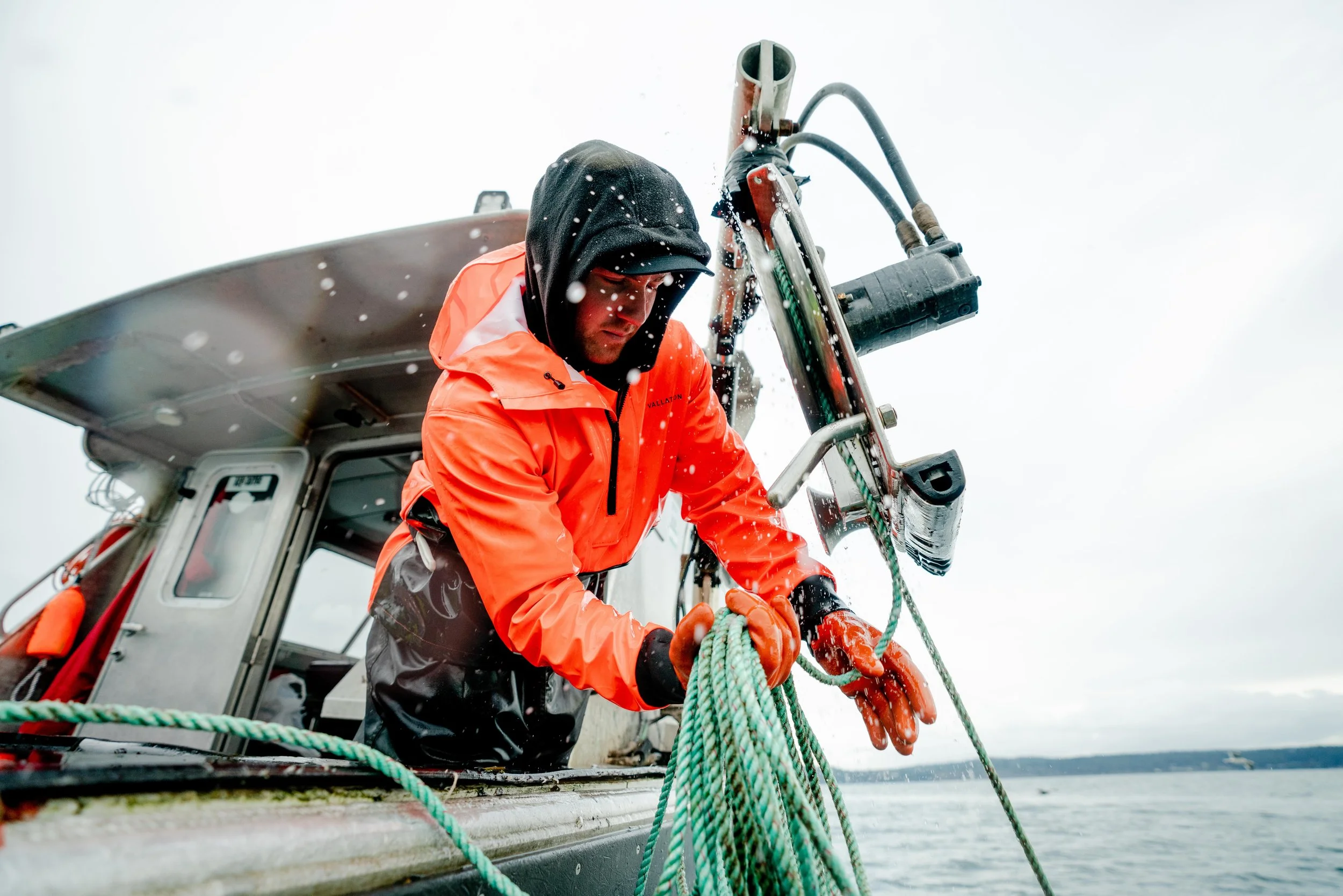 A man in orange waterproof gear and black hoodie handling green ropes on a boat in a rainy setting.
