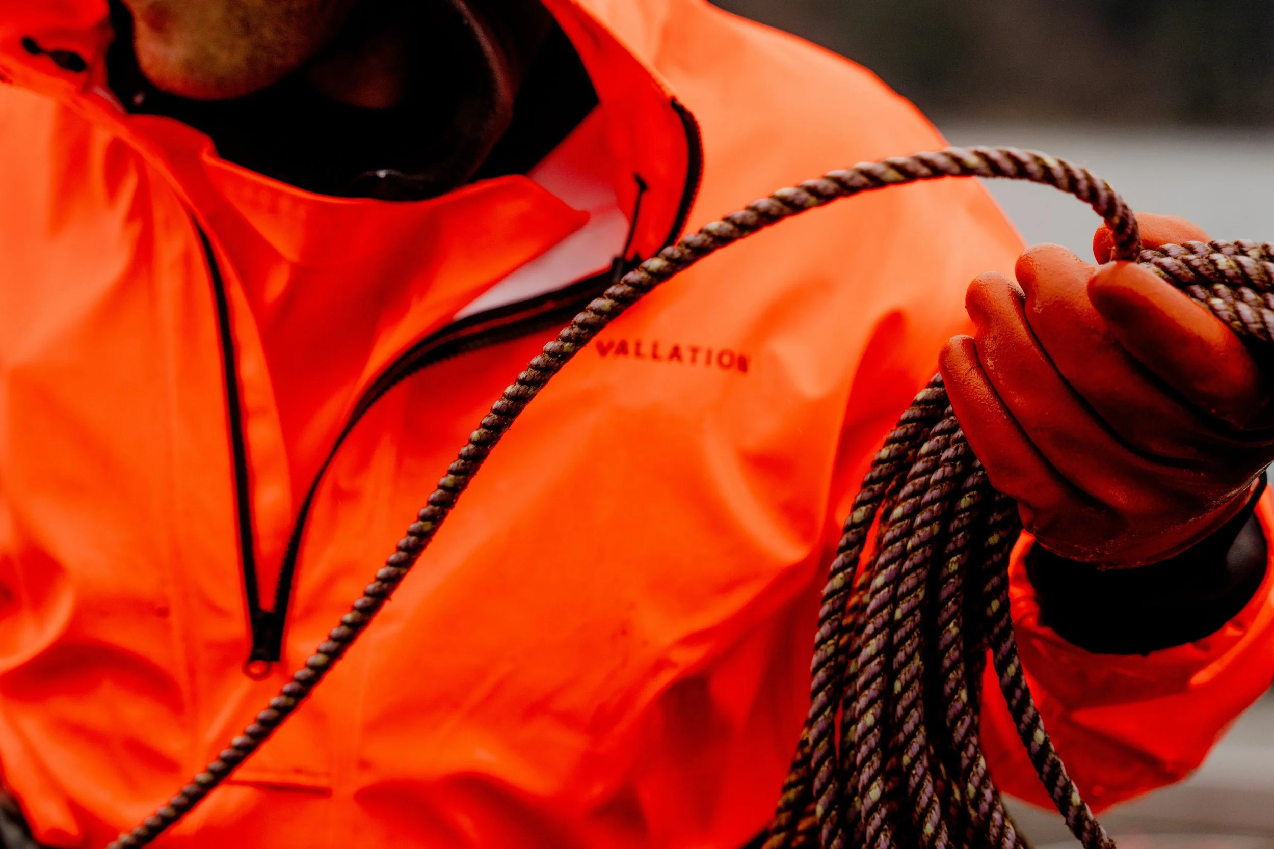 Close-up of a person wearing an orange Vallation jacket holding a coil of rope and wearing gloves.