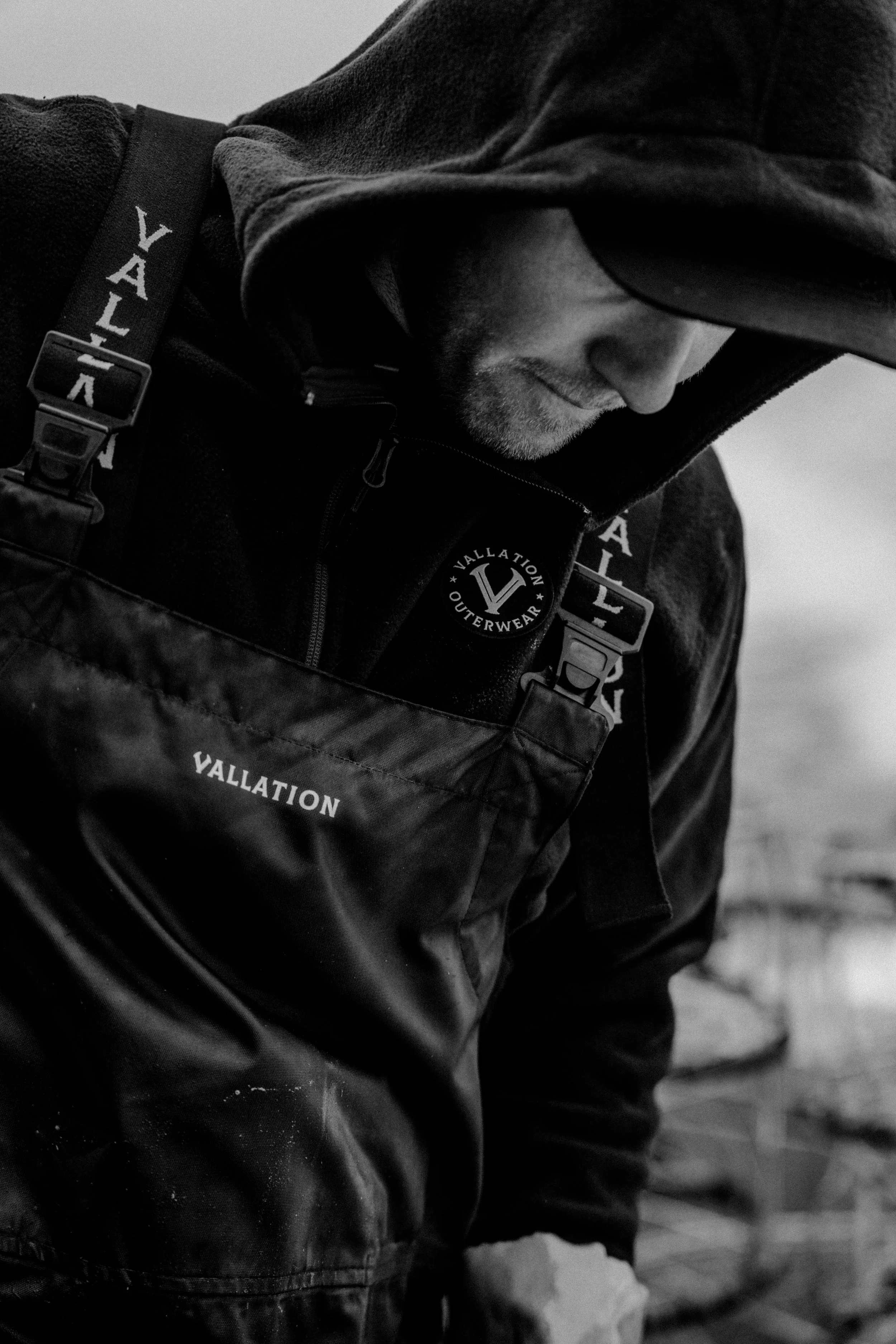 Close-up black and white photo of a man wearing a hooded jacket, with the hood over his head, looking down. The jacket and gear display the brand name 'Vallation'.