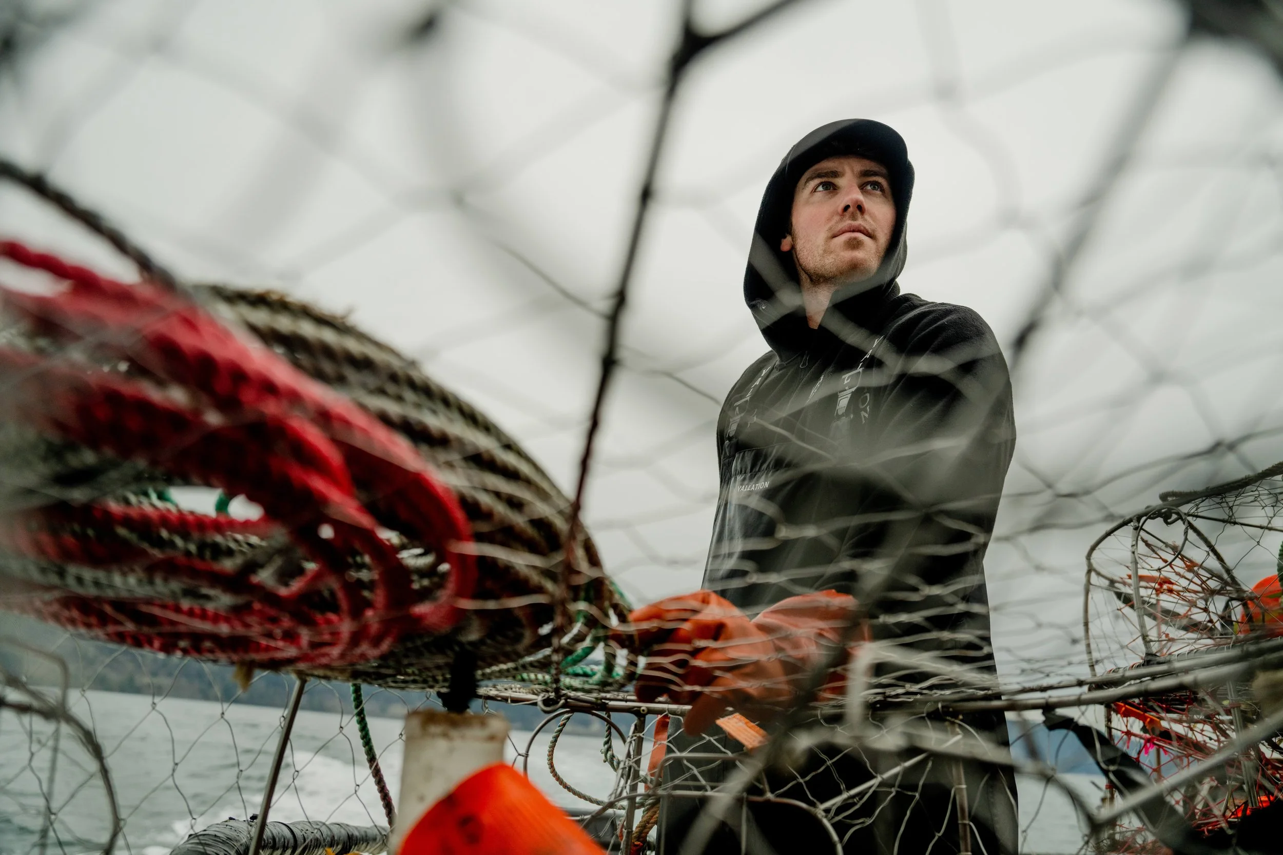 A man in a black hoodie and orange gloves standing on a boat surrounded by fishing nets and equipment, with a body of water in the background and an overcast sky.