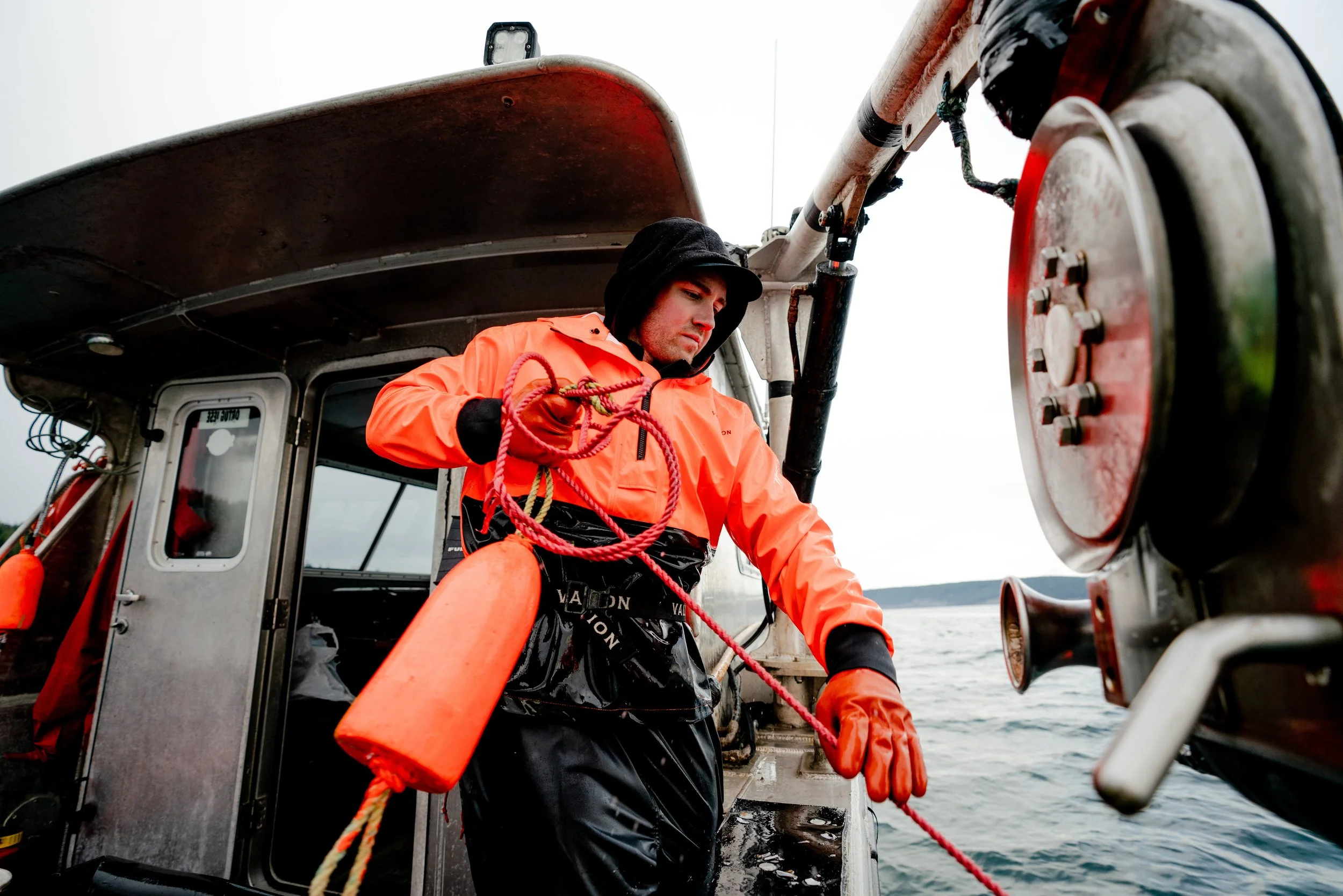 A man in an orange waterproof jacket and gloves on a boat, handling a red rope near a large winch, with water and a distant shoreline in the background.