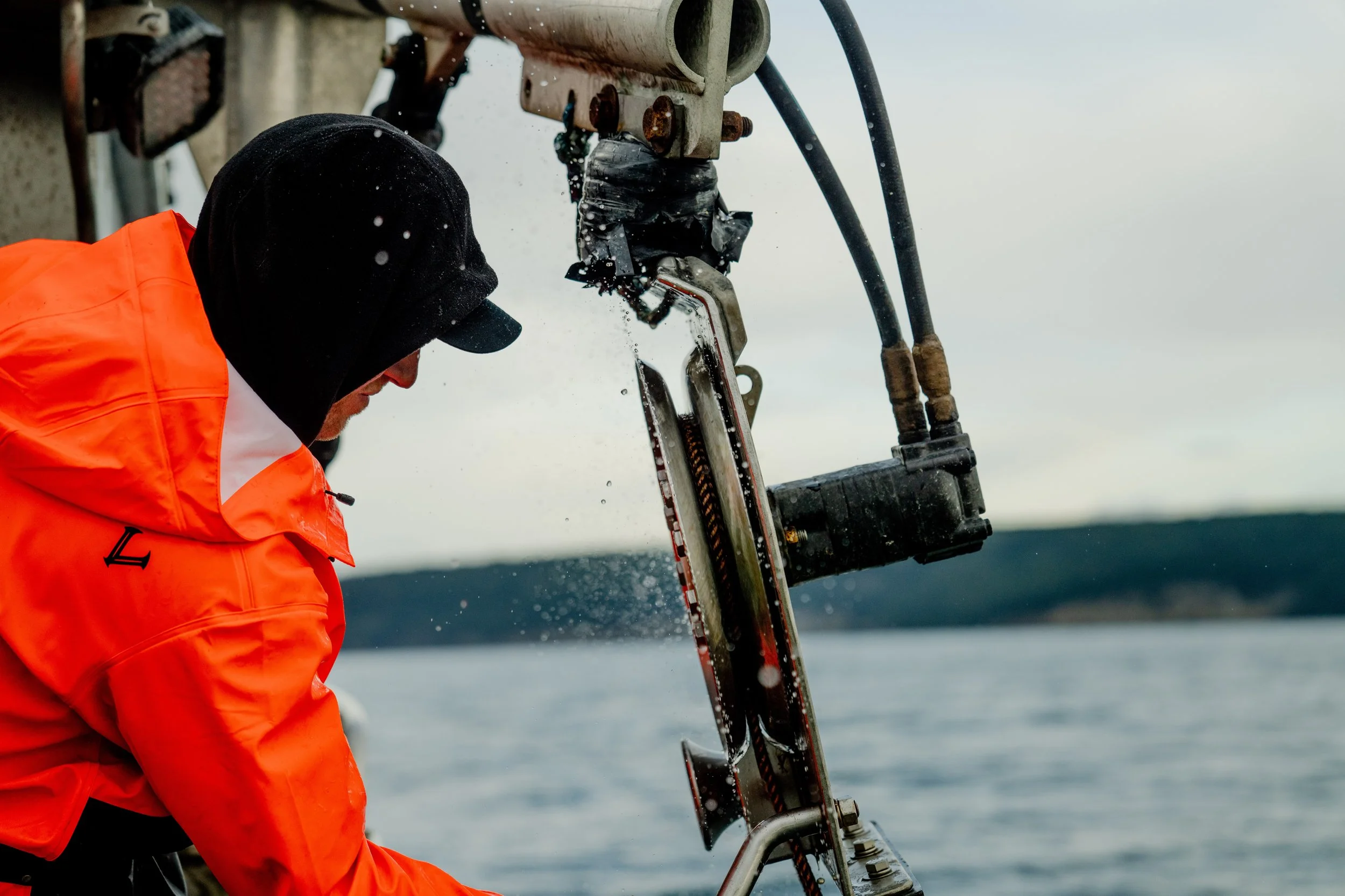 A person wearing a black hood and orange waterproof jacket operating a mini submarine or underwater drone near water.