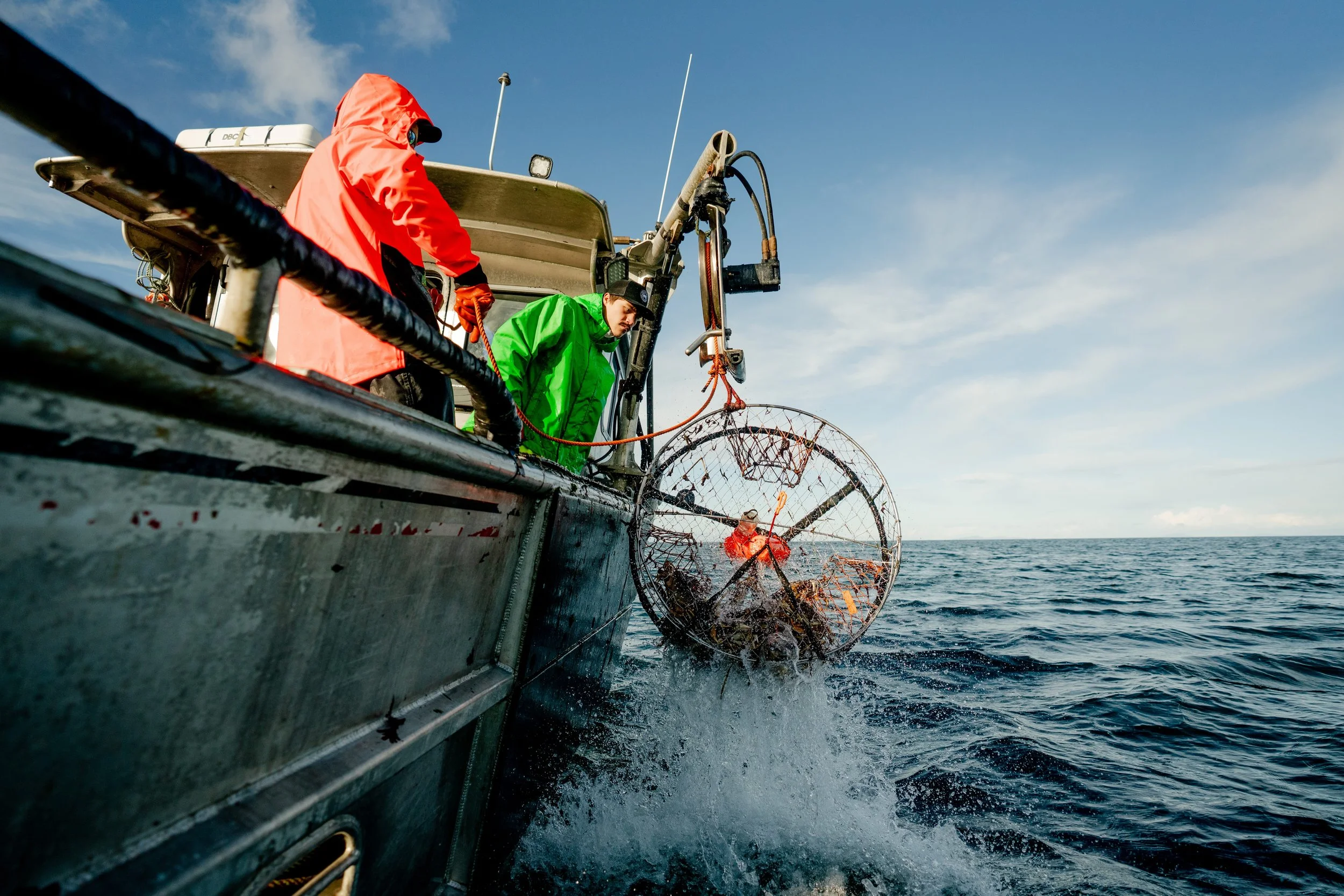 Two scientists in waterproof jackets aboard a research vessel lowering a net into the ocean for marine research on a clear day.
