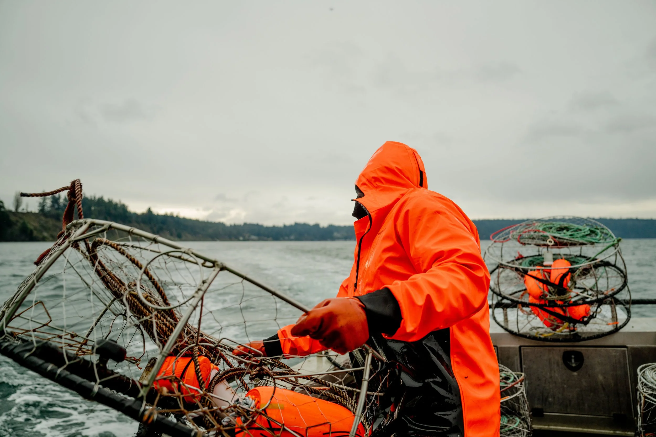 Person wearing an orange raincoat and black gloves onboard a boat, handling fishing traps, with overcast skies and water in the background.