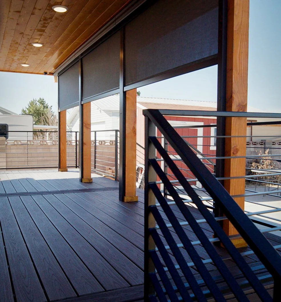 View of a residential balcony with wooden flooring, wooden support beams, black metal railing, and outdoor roller blinds partially rolled down, with a neighboring house and trees visible in the background.