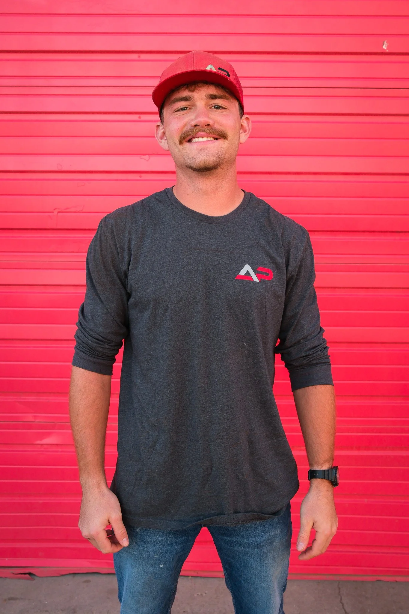 A young man wearing a red baseball cap and matching red t-shirt with a logo, standing in front of a red panel background, smiling at the camera.