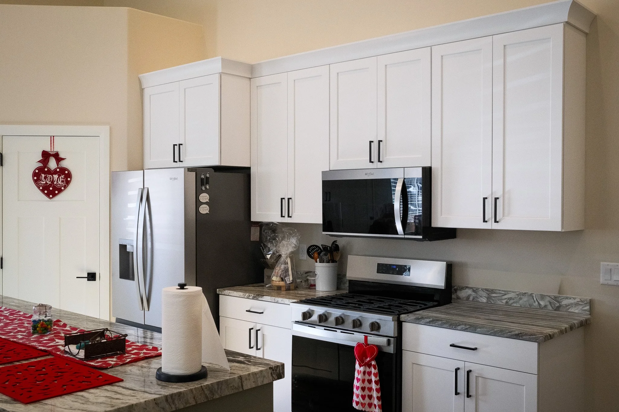 Kitchen with white cabinets, black appliances, a stainless steel refrigerator, and a marble countertop. Valentine's Day decorations include a red heart ornament on the oven handle, a red heart-shaped oven mitt, and a red and white table runner with Christmas themed items on the table.