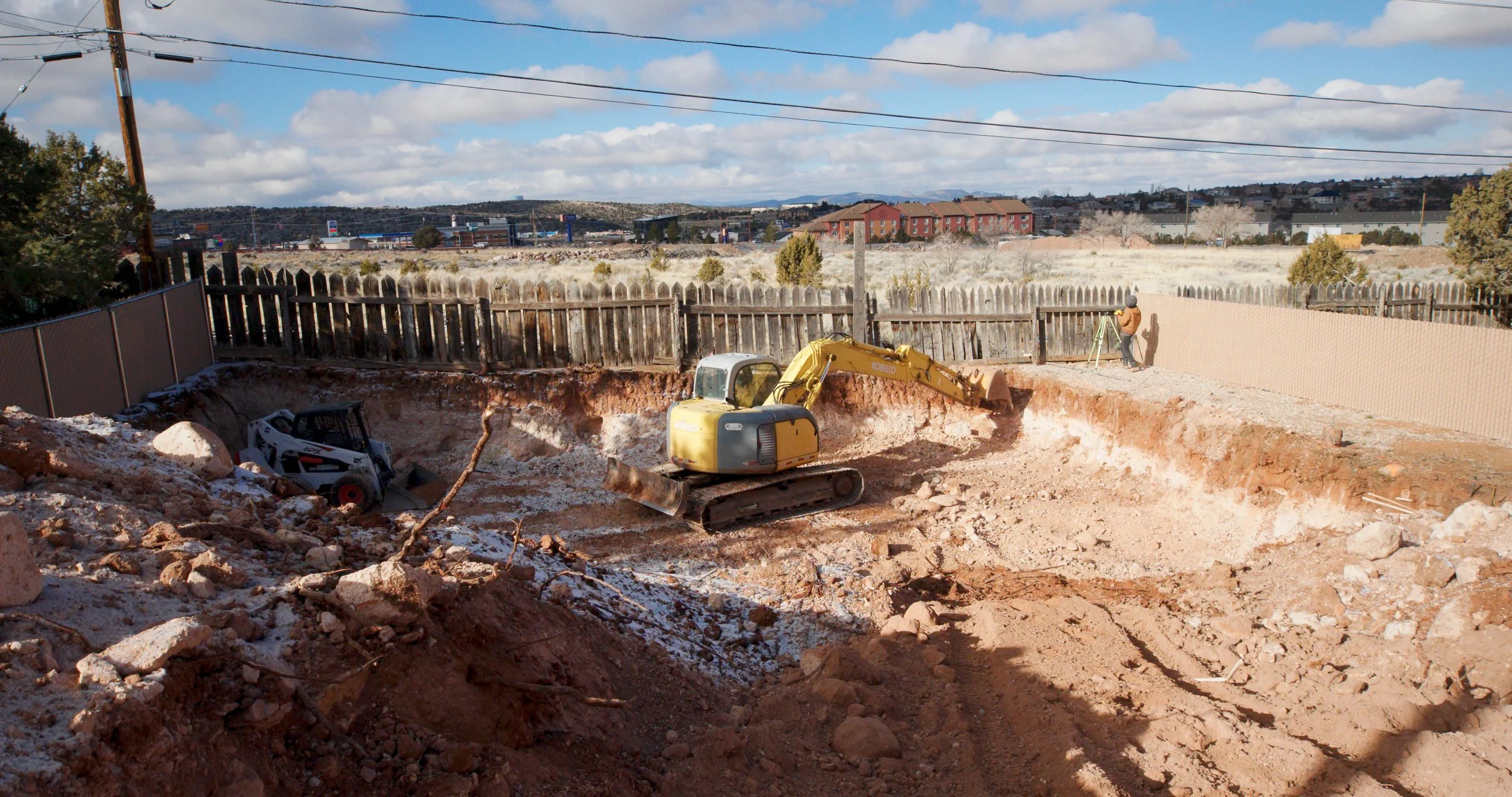 Construction site with excavator digging in a dirt lot, surrounded by a wooden fence, with a person surveying with a tripod and level nearby, under a partly cloudy sky.