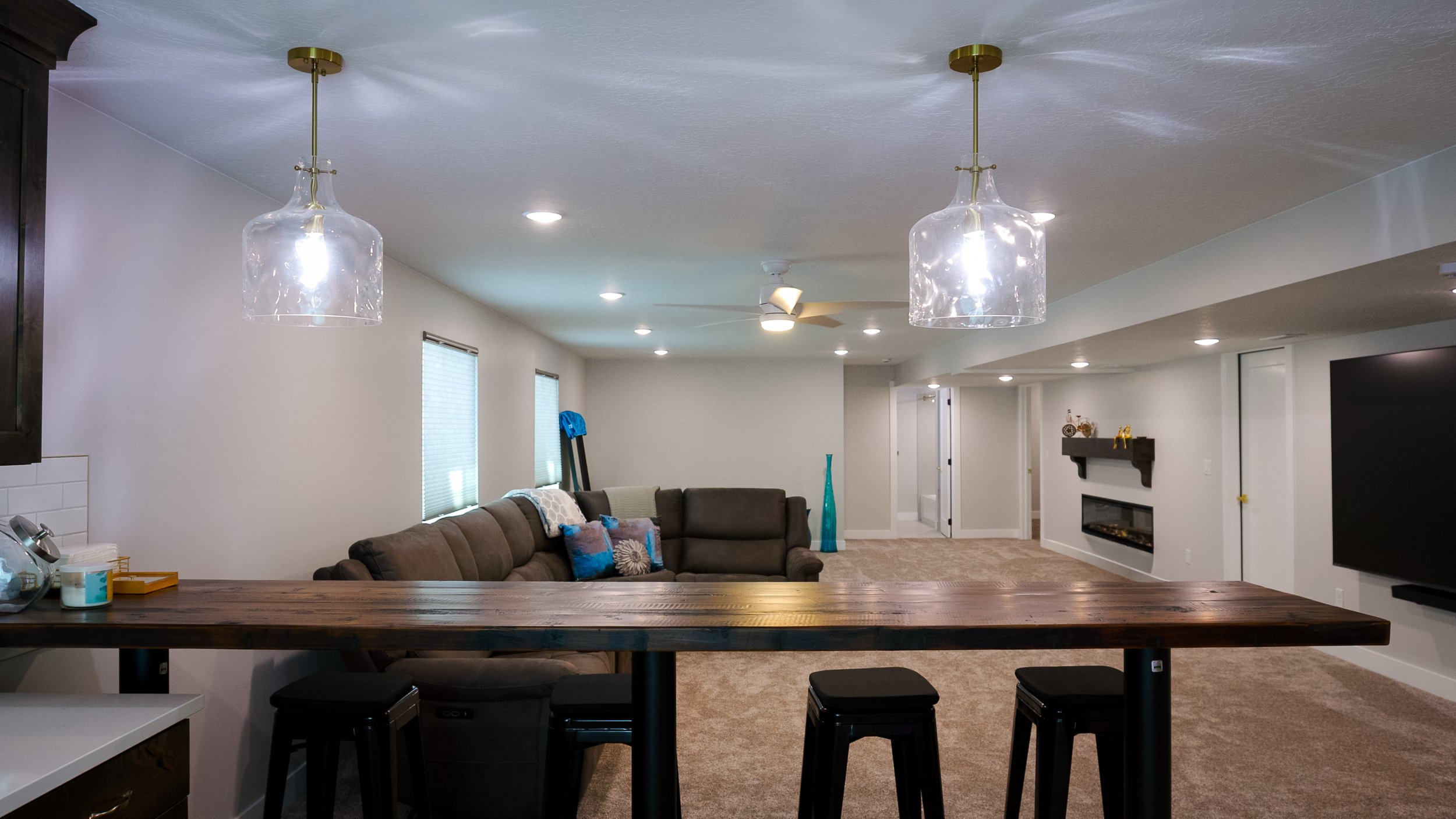 Open-concept living room with ceiling fans, recessed lighting, a beige carpet, a fireplace with a black mantel, a large sectional sofa with patterned pillows, and a dining area with a wooden table and black stools.