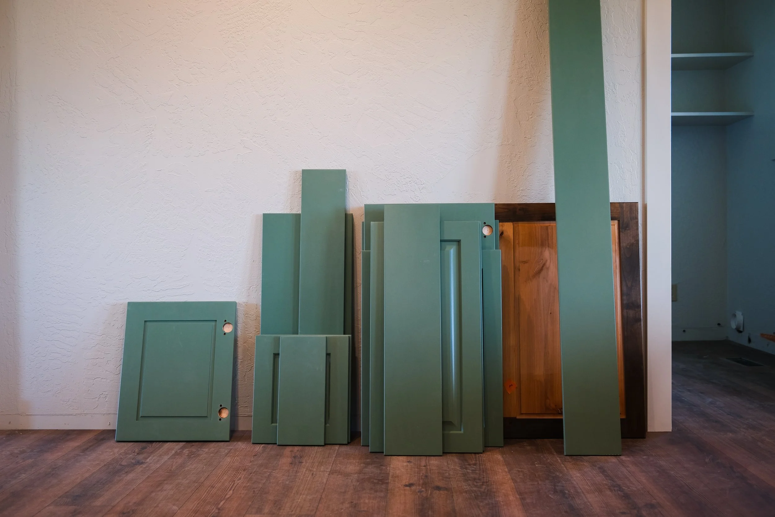 Various green painted cabinet doors and panels leaning against a wall in a room with wooden floor, with a partial view of more cabinets and shelves on the right.