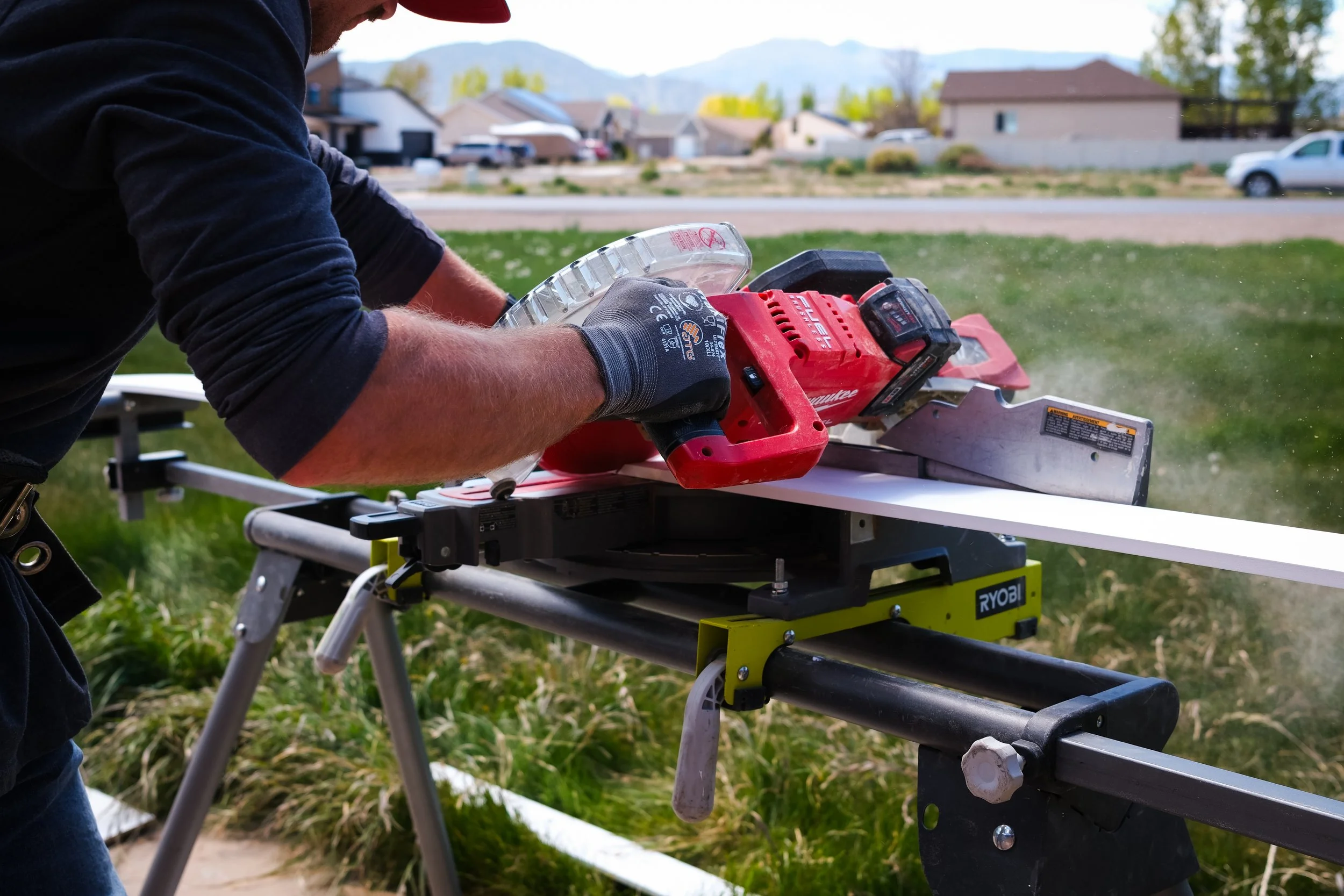 A person wearing a black long sleeve shirt and gloves using a red cordless circular saw on a workbench outside in a residential area with houses, grass, and mountains in the background.