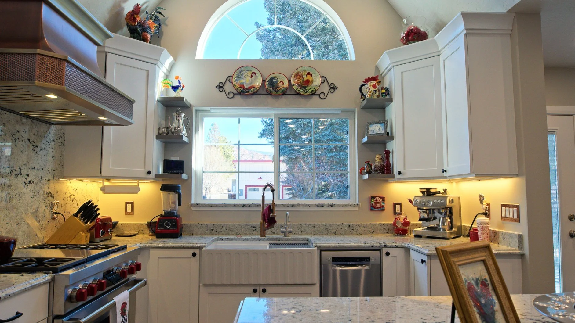 A kitchen with white cabinets, granite countertops, and a large window with an arched top. The window shows trees and a house outside. The countertop has a red blender, knife block, coffee maker, and various decorative items, including rooster figuri