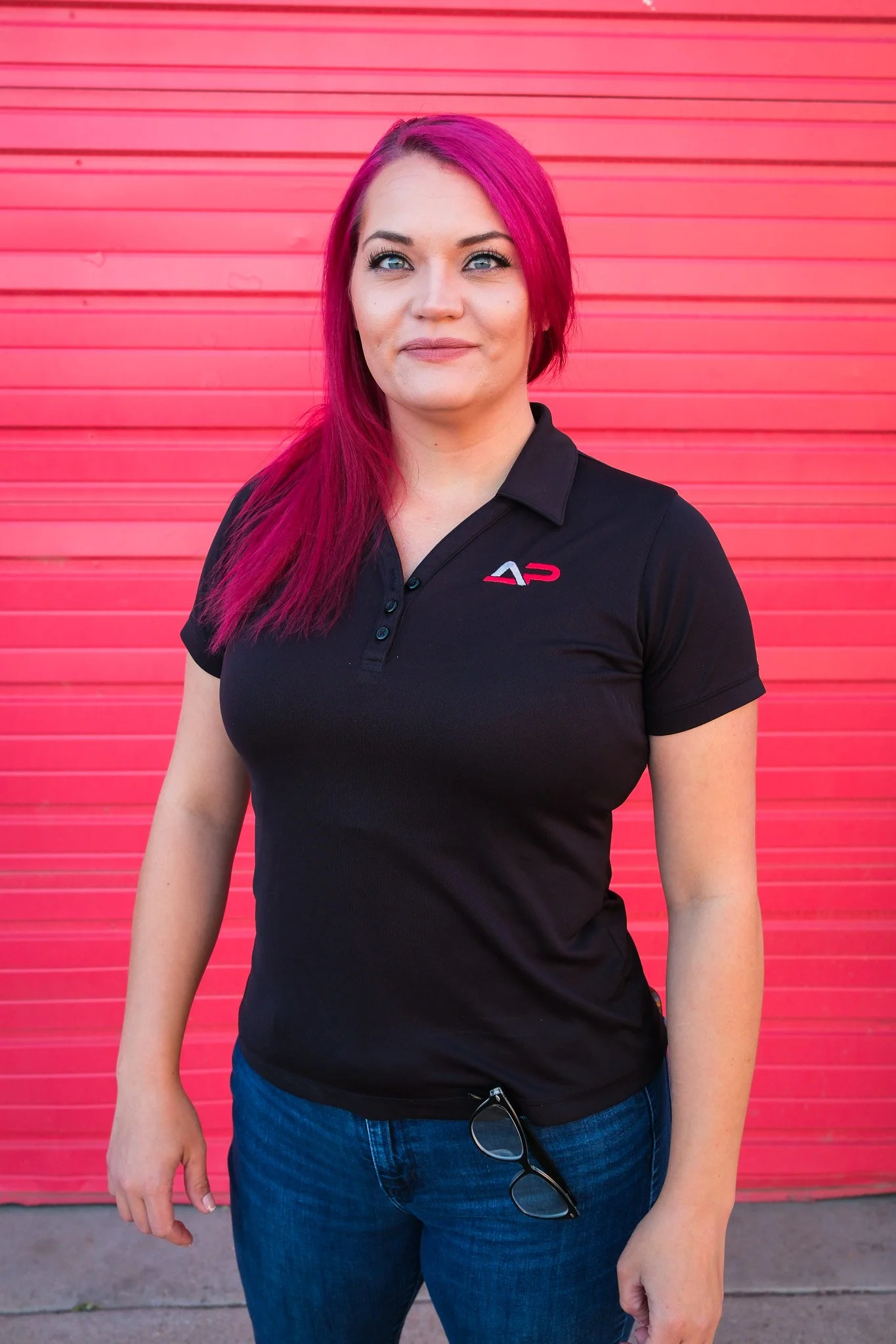 A woman with dark hair standing in front of a red corrugated metal wall, wearing a red polo shirt with a logo and black pants.
