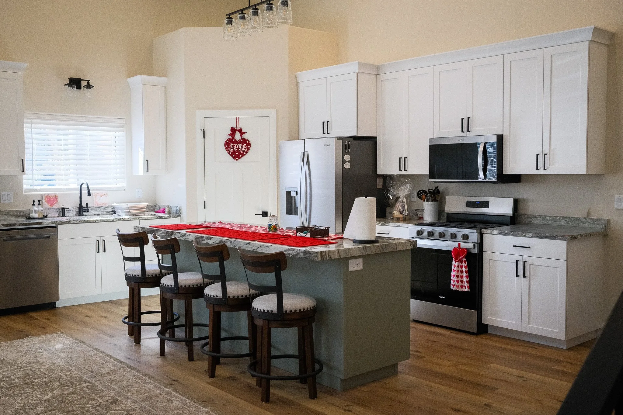 A modern white kitchen with a marble island, four chairs, and a Valentine's Day decoration on the wall.