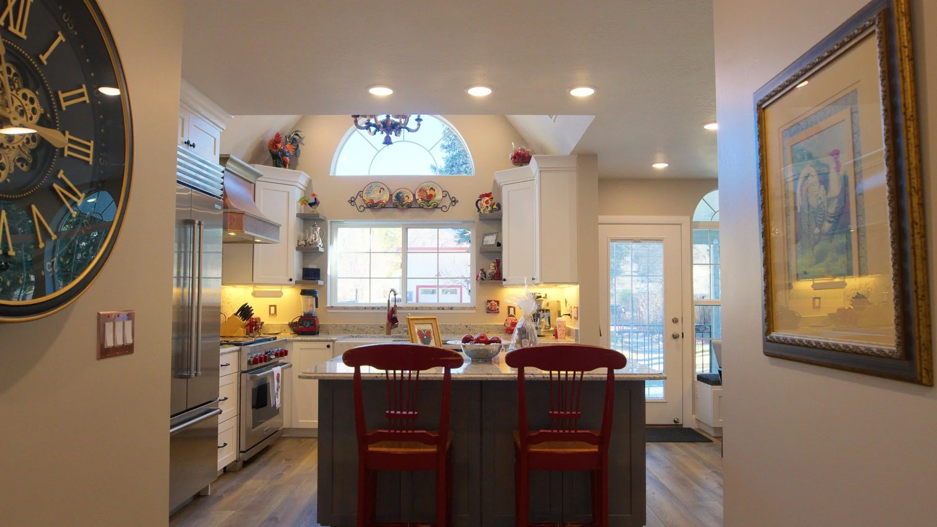 View of a bright kitchen with a large window, white cabinets, a stainless steel refrigerator, a kitchen island with red chairs, and decorative plates above the window.