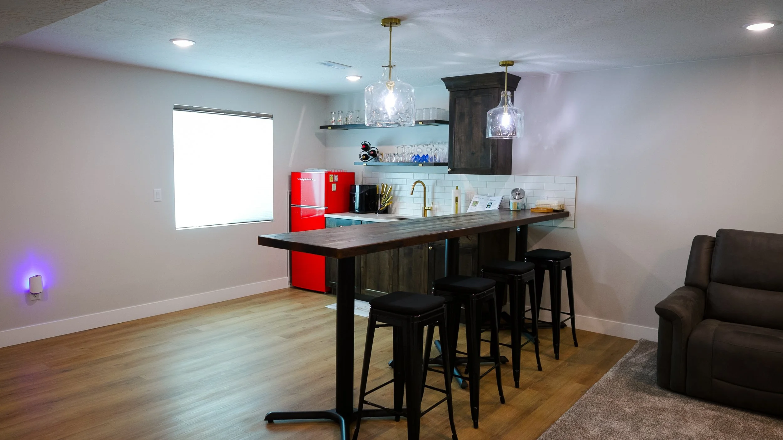 Interior view of a modern kitchen with a bar-height wooden counter, black bar stools, and a red mini fridge, with shelves holding glasses and a wine rack, illuminated by ceiling lights and pendant lamps.
