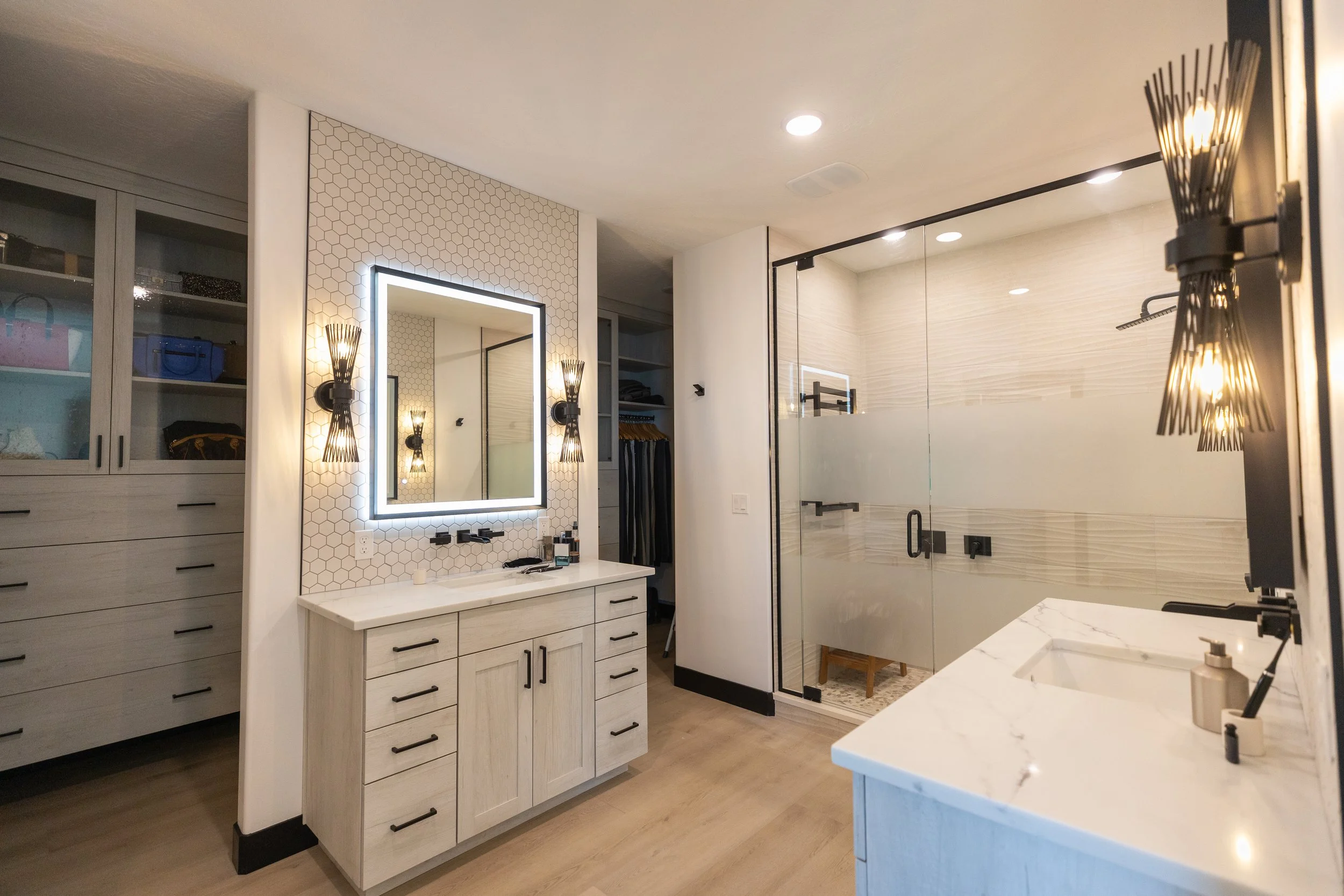 Modern bathroom featuring a white vanity with black hardware, a large backlit mirror, hexagonal wall tiles, black wall-mounted light fixtures, a glass-enclosed shower with beige tile walls, and open closet shelves with bags and clothing.